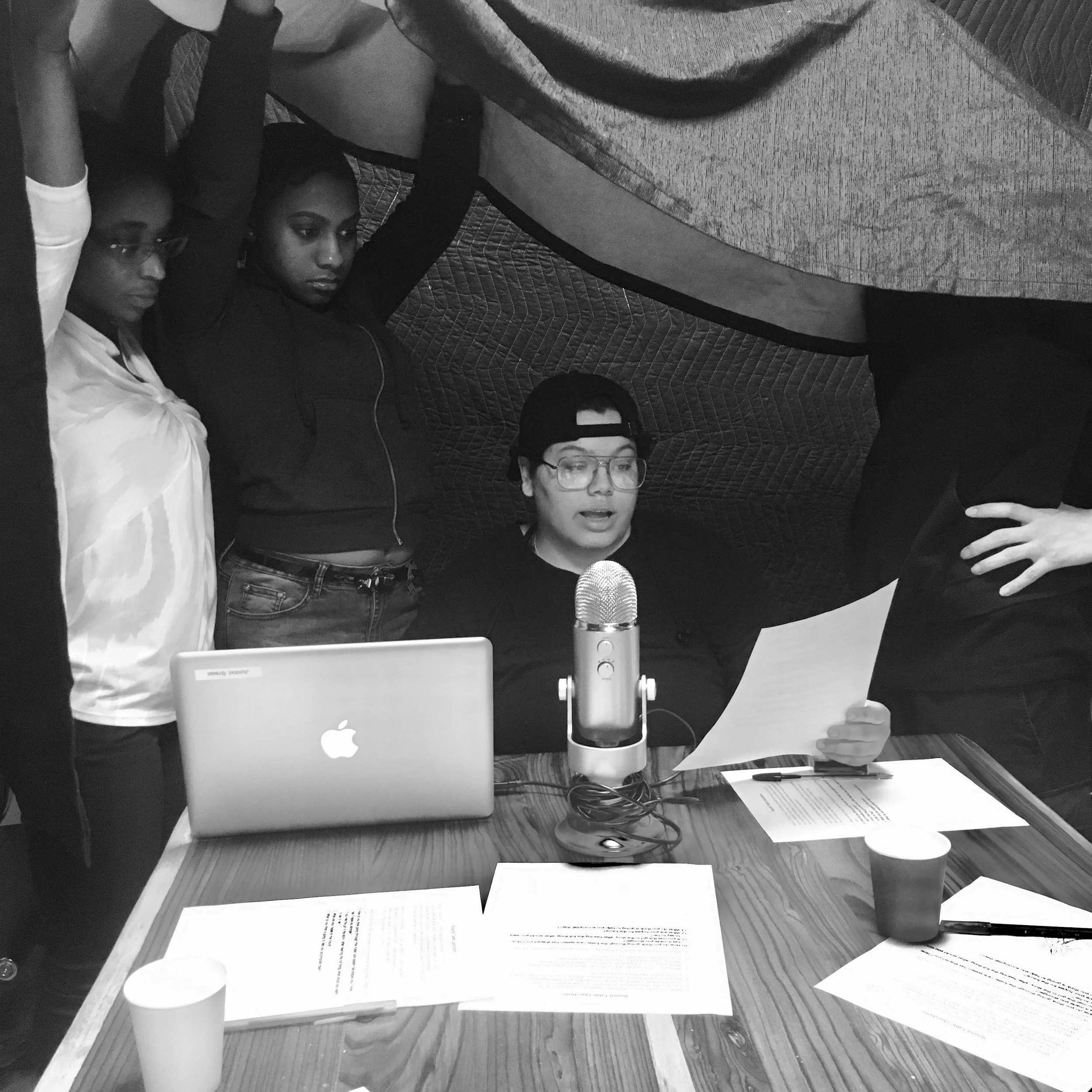 Four young people are gathered around a table with laptops, papers, and cups, with one person seated and speaking into a microphone, while the others stand or lean nearby, in an indoor setting.