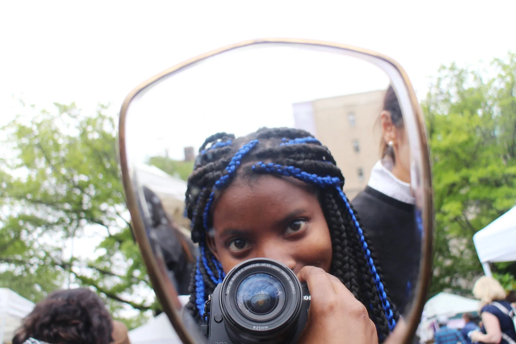 A woman with braided hair and blue hair extensions is taking a selfie with a camera, reflected in a car's side mirror. She is outdoors with trees and tents in the background.