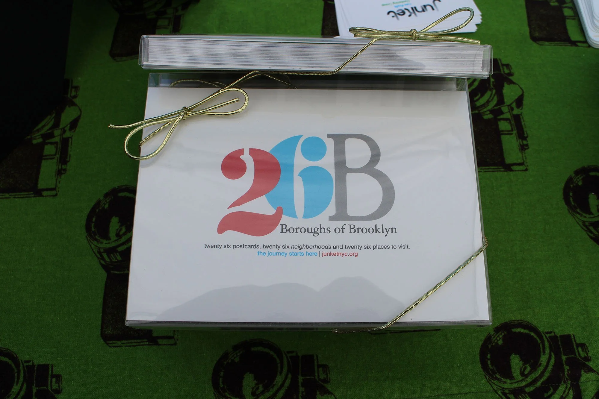 A brochure titled '26 Boroughs of Brooklyn' features with colorful text and a subtitle about visiting neighborhoods and places in Brooklyn, resting on a green tablecloth with a black camera pattern.