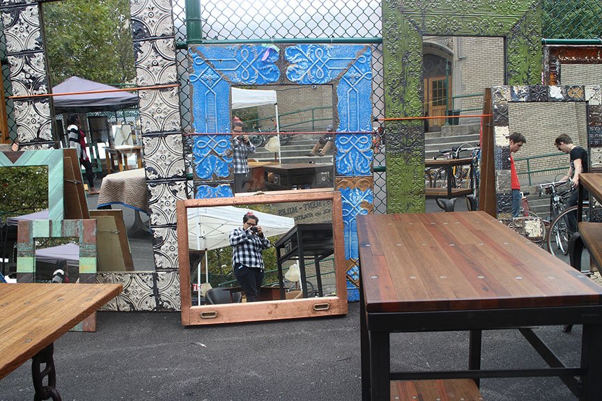 A collection of various decorative mirrors and frames leaning against a wall outdoors, with reflections of people and trees visible in some of the mirrors.