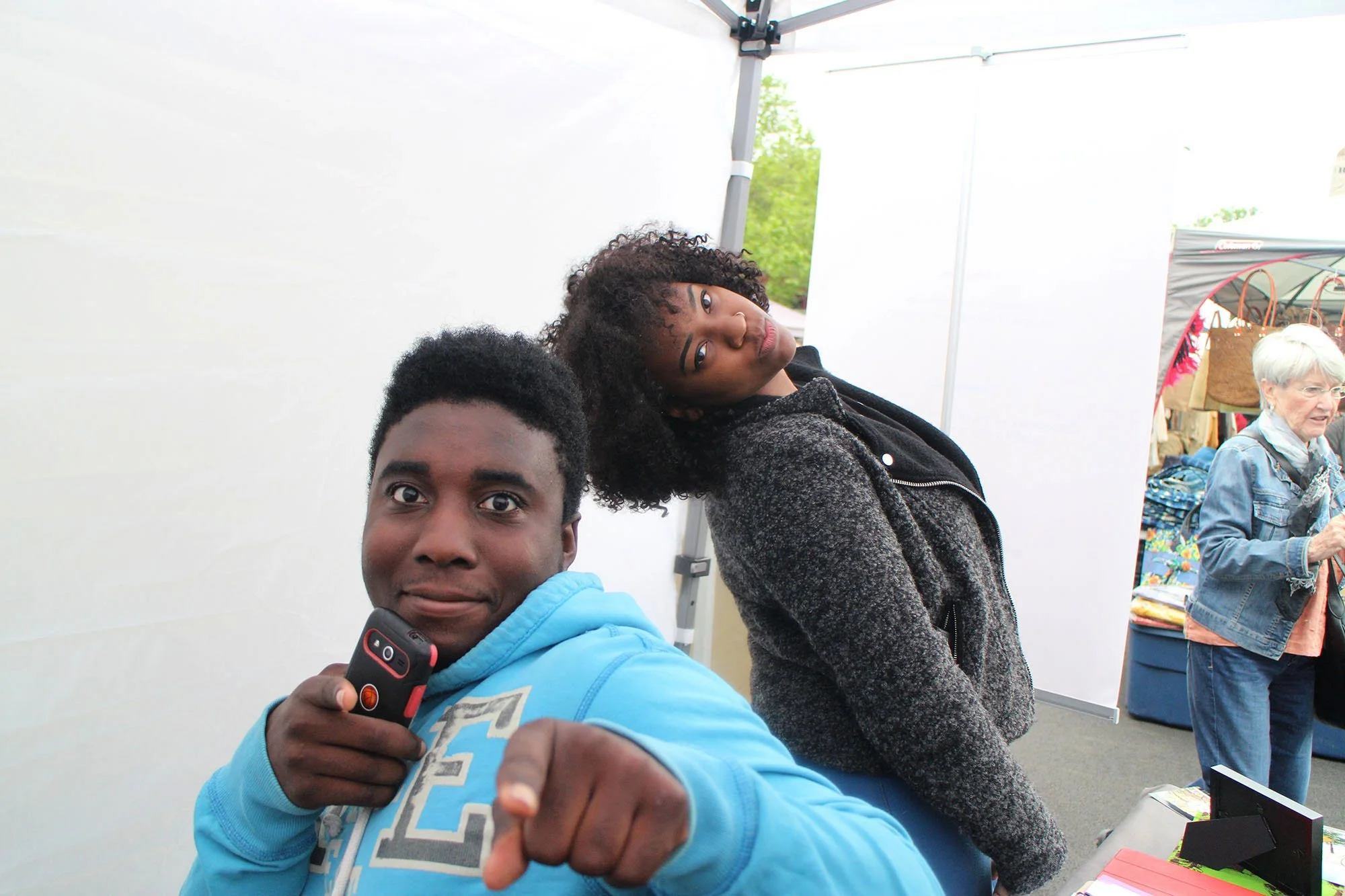 Two young people, a man and a woman, are posing playfully at an outdoor event or market. The man is holding a camera and pointing at the viewer, while the woman leans her head back, tilting to the side. In the background, an older woman is browsing i