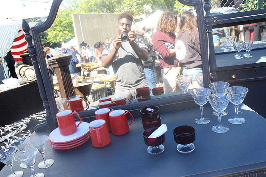 Reflection of a young man taking a photo with a camera in a mirror at an outdoor market with various glassware and tableware on display.