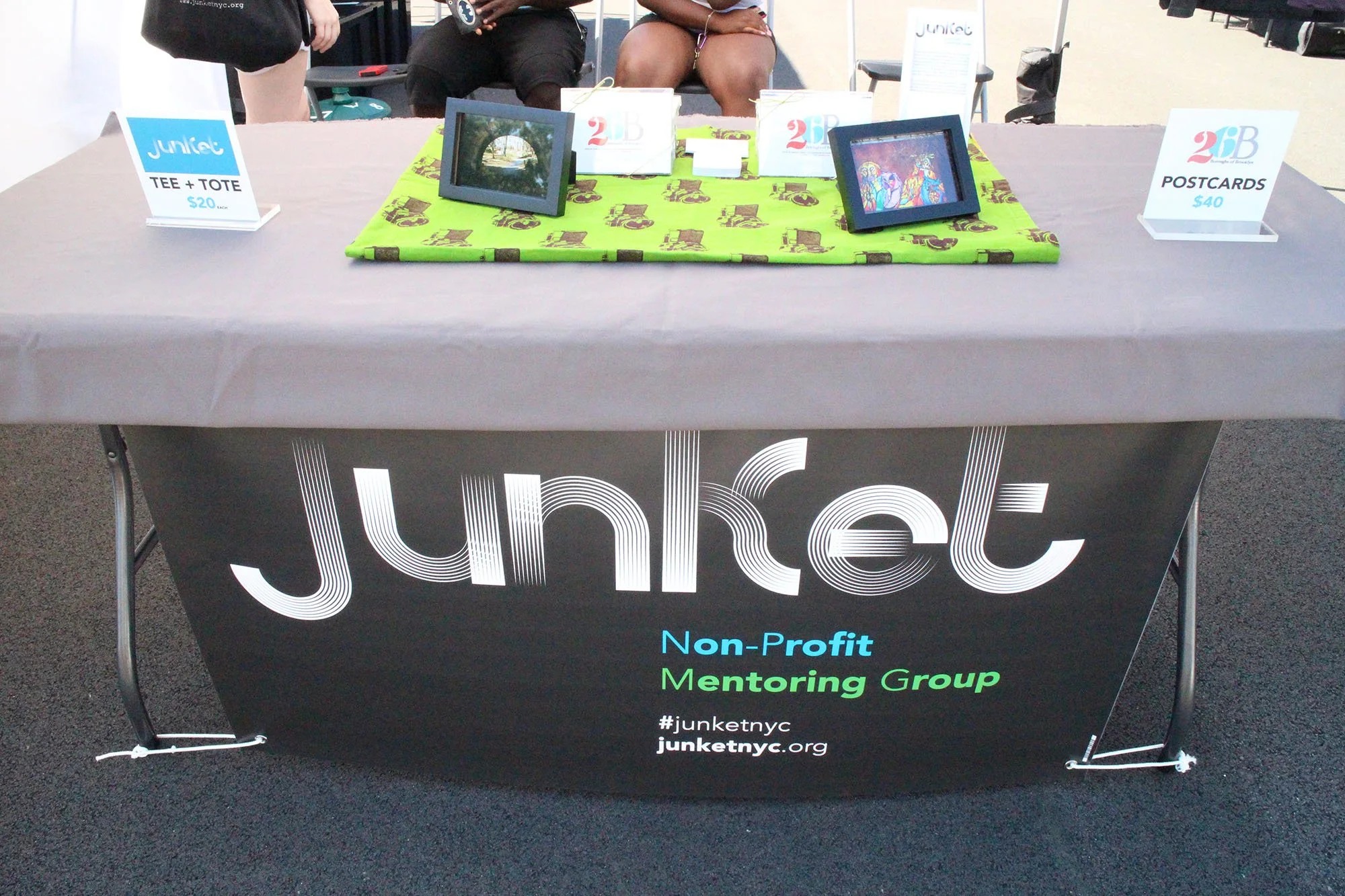 Table with merchandise and signage for Junket, a non-profit mentoring group, with signs advertising T-shirts + totes for $20 and postcards for $40.