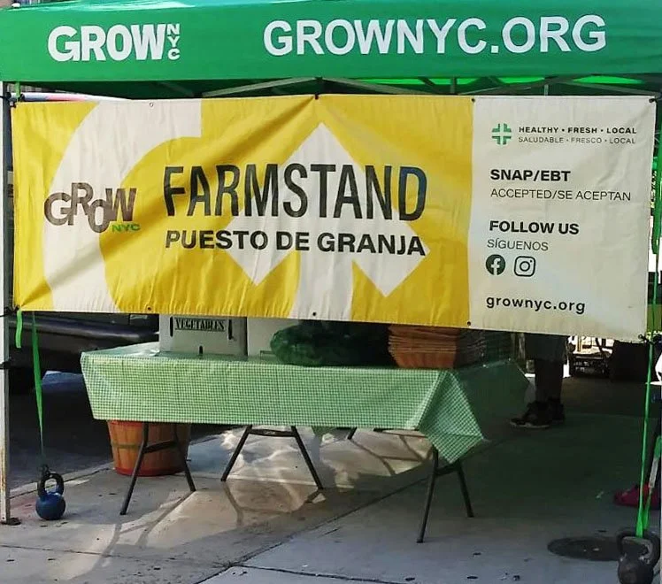 A green tent with white and yellow banners advertising a farmer's market stall for Grow NYC. There is a table covered with a green checkered cloth underneath the tent.
