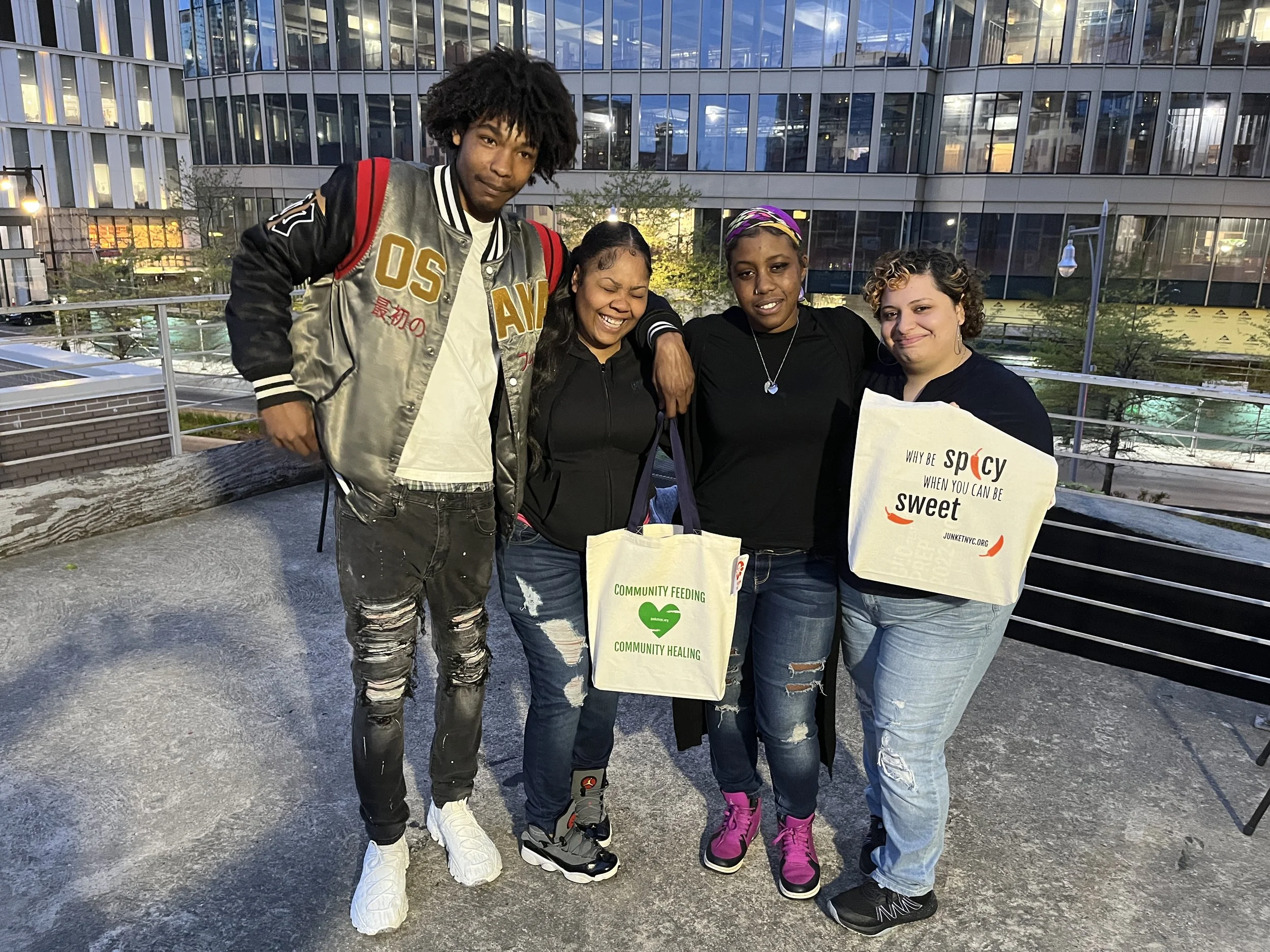 Four people standing together on a rooftop, smiling. They are holding reusable bags with messages about community feeding, healing, and spice. The background shows a cityscape with modern glass buildings.