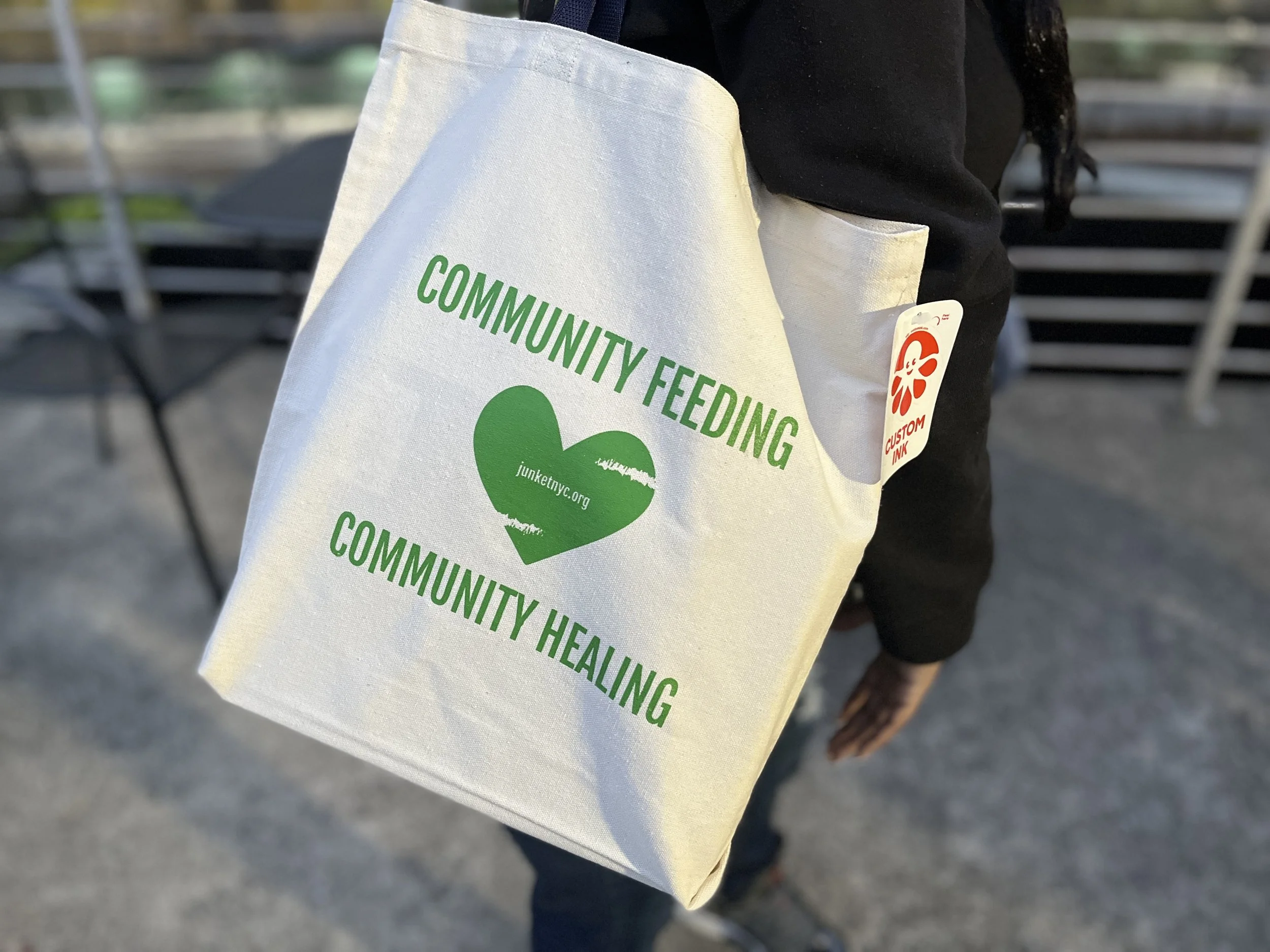 Person holding a white tote bag with green text that reads 'Community Feeding' and 'Community Healing,' with a green heart and a website URL inside it.