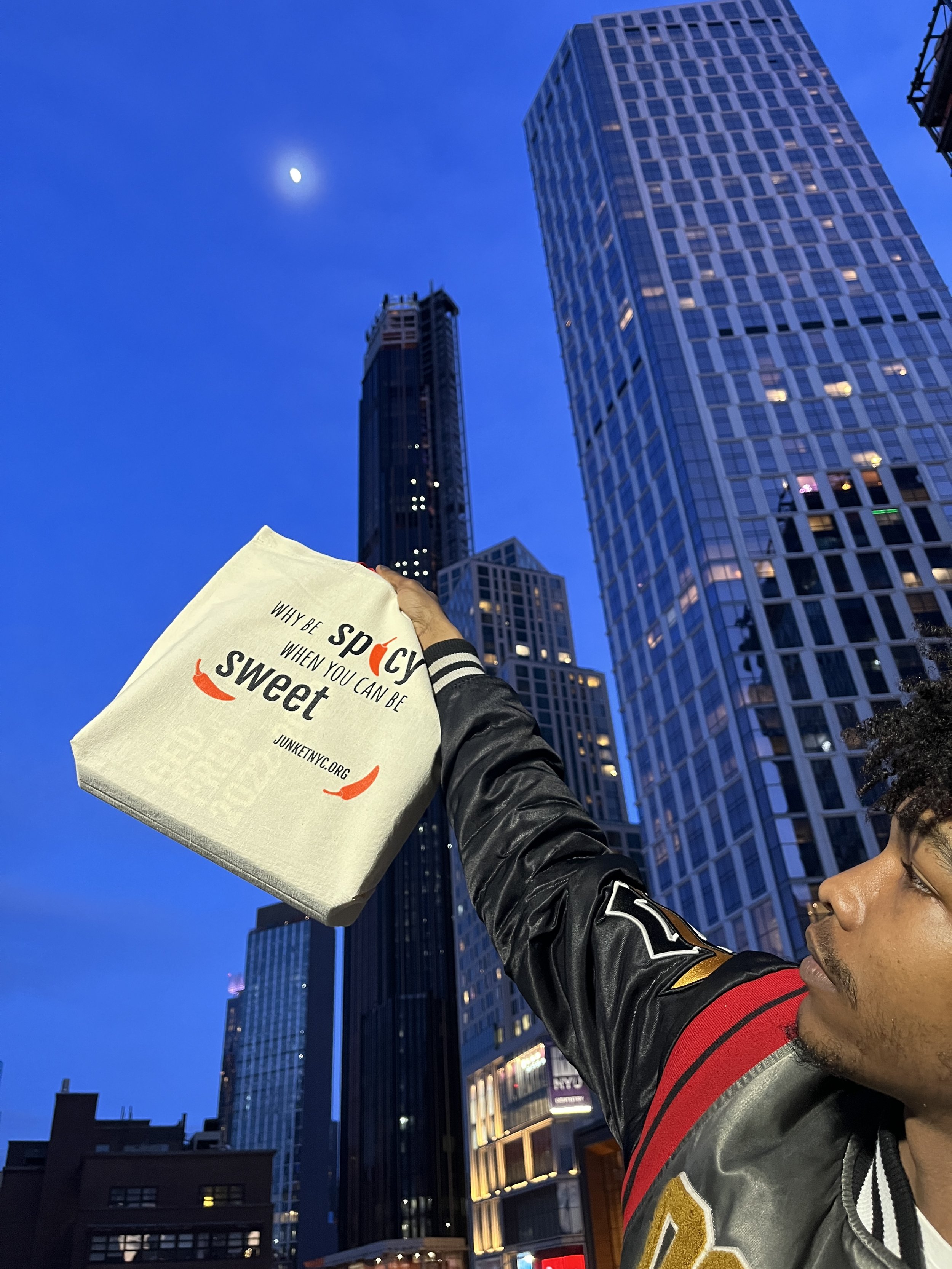 A man holding a white tote bag with the words 'Why be spicy when you can be sweet' in front of tall city skyscrapers at dusk, with a visible moon in the sky.