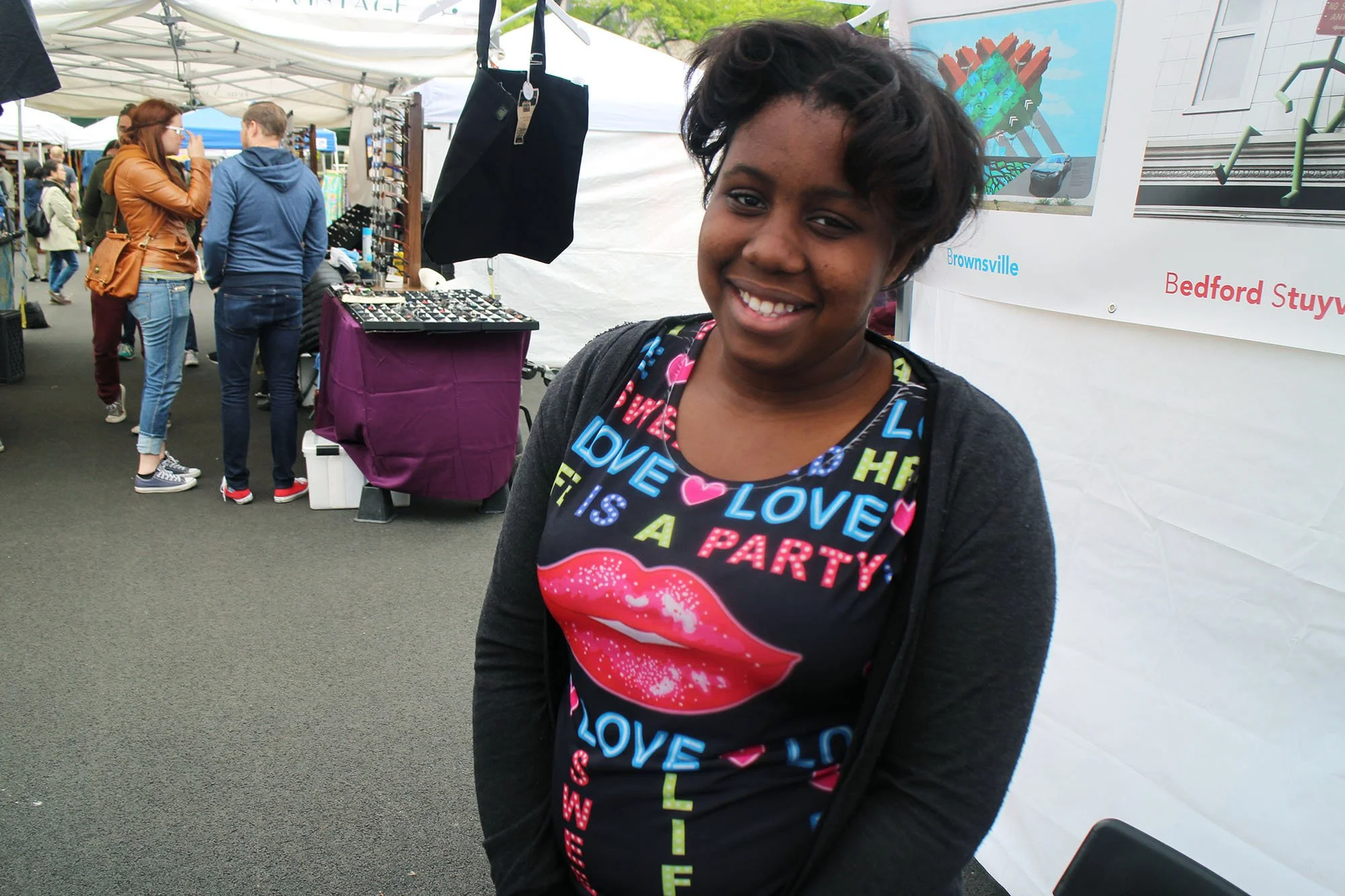 Young woman smiling at an outdoor market, wearing a dark shirt with colorful text and lips design, with vendors and shoppers in the background.