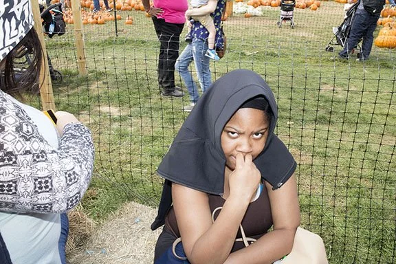 Woman in a black headscarf and brown top standing in front of a pumpkin patch, with her hand near her nose, and others in the background.