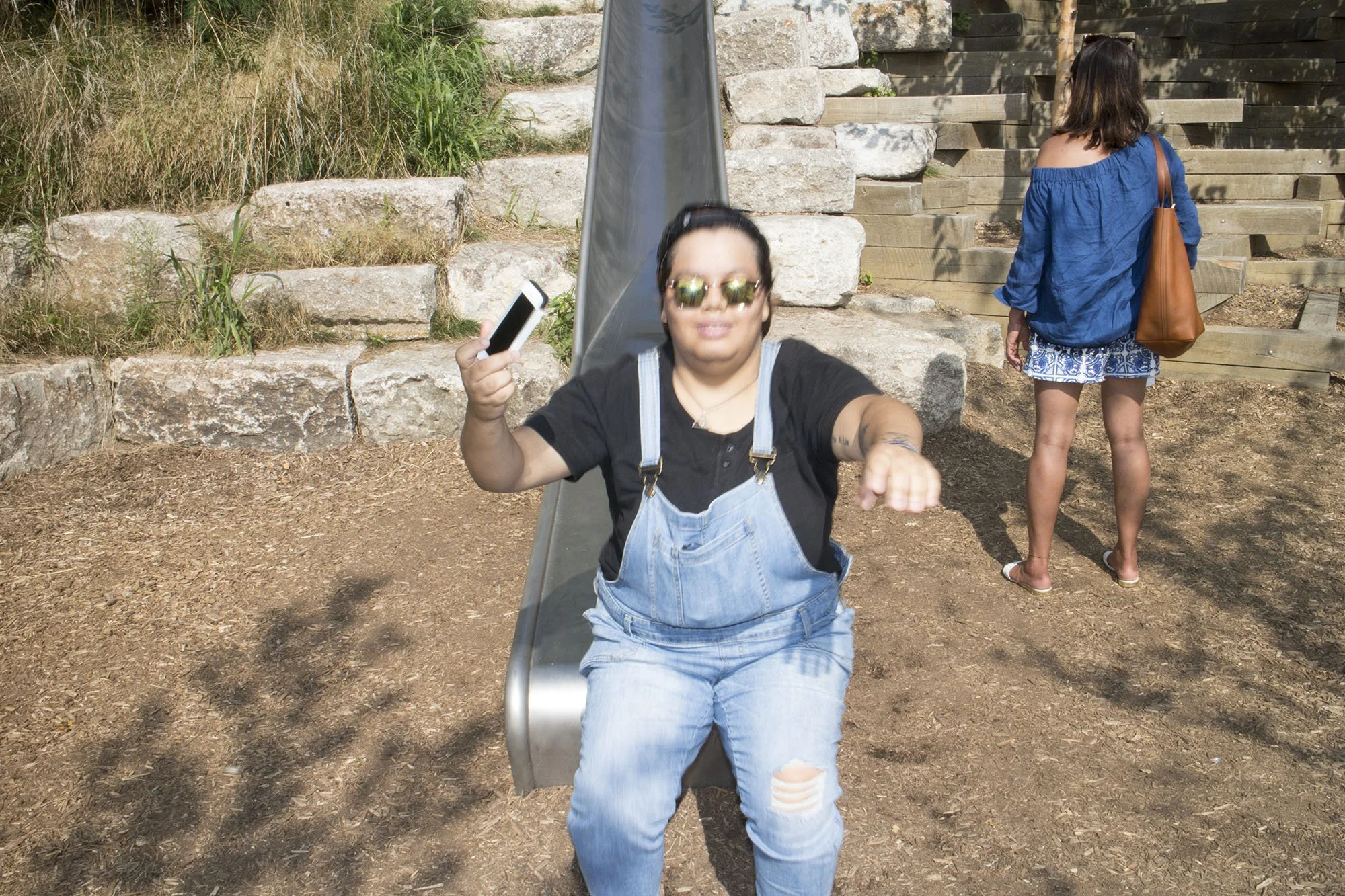 A woman sitting on a slide, holding a smartphone, at an outdoor playground, with a young girl in a blue jacket and patterned shorts standing nearby with her back to the camera.