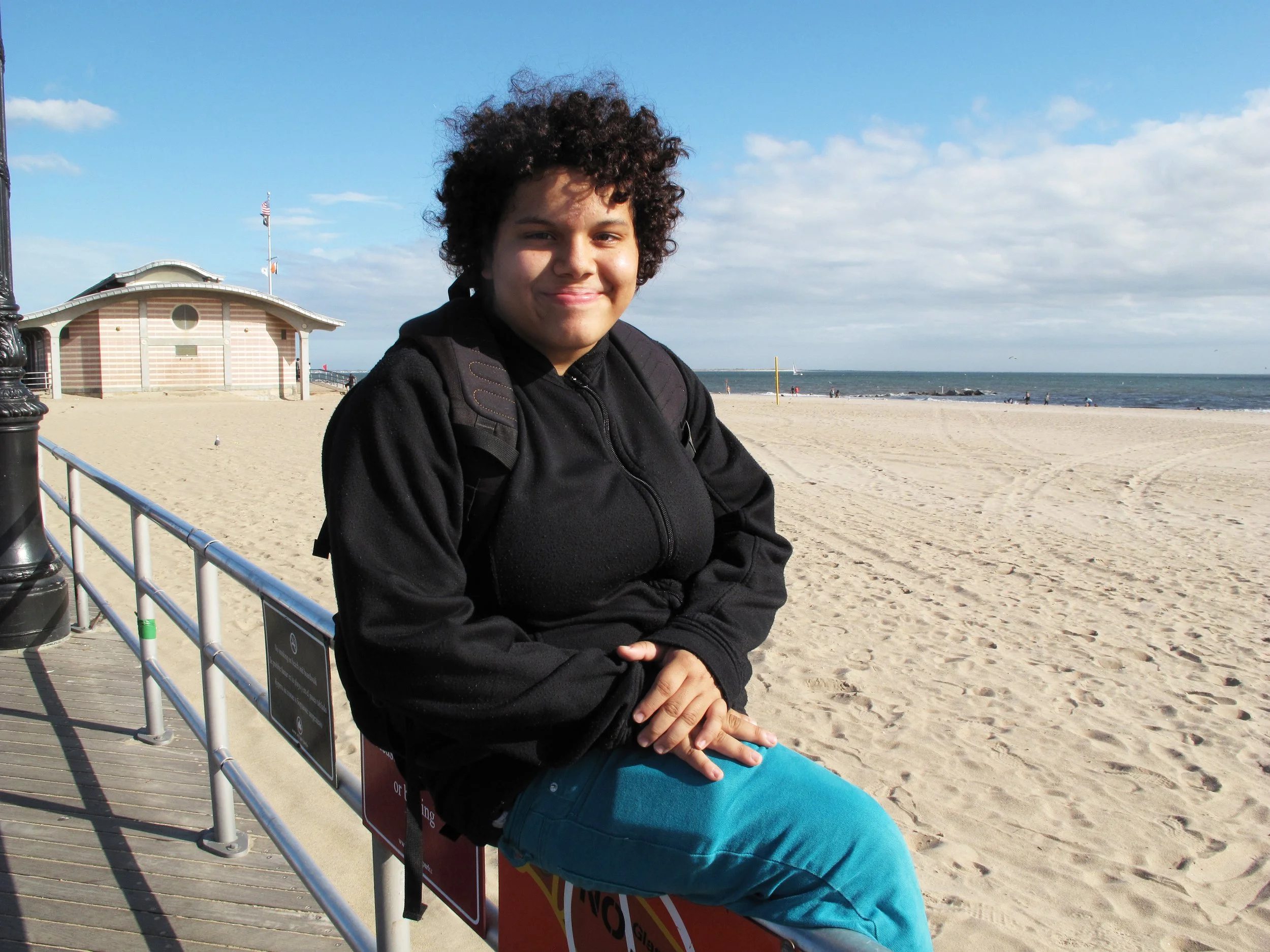 A woman with curly hair sitting on a bench at the beach, smiling. She is wearing a black jacket, blue pants, and has a backpack. The sandy beach, ocean, and sky with clouds are visible in the background.
