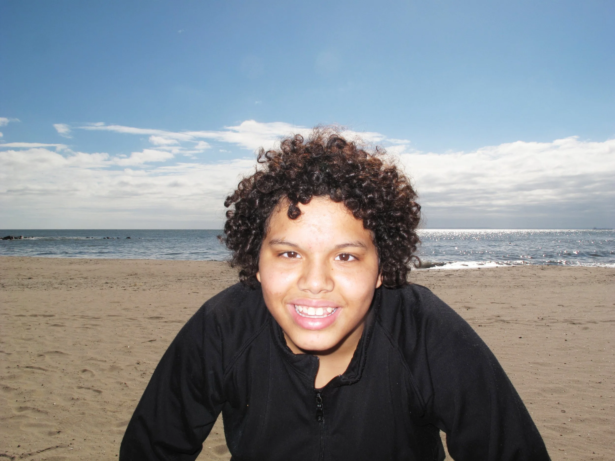 A smiling child with curly hair on a sandy beach near the ocean under a partly cloudy sky.