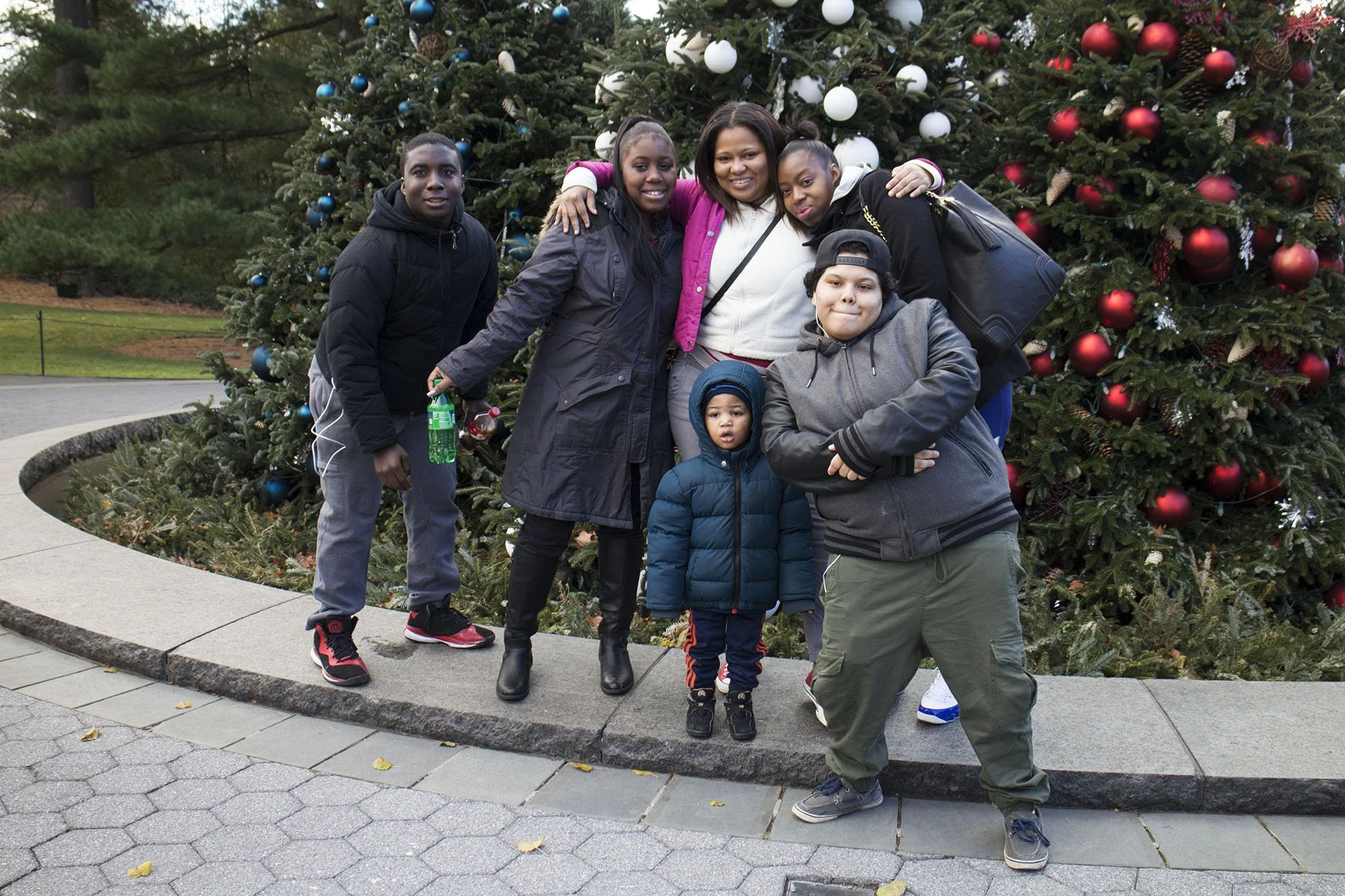 A group of six people, including children and adults, posing in front of a large decorated Christmas tree outdoors.