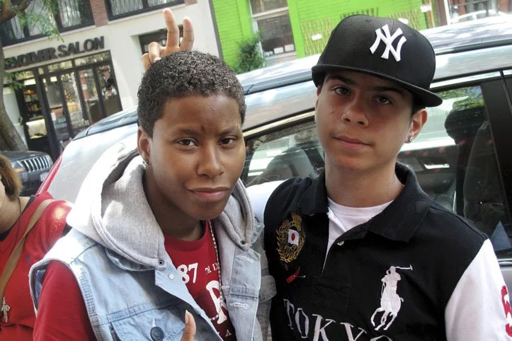 Two young males standing outdoors in front of a car; one is making a hand gesture and the other is looking at the camera. They are dressed casually, one wearing a baseball cap and the other a hoodie.