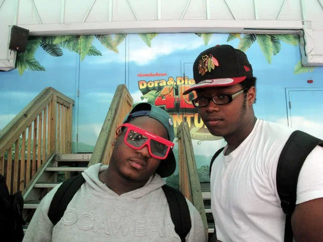 Two young men wearing backpacks and sunglasses standing in front of a printed backdrop for "Dora & Die 40 Adventure" at an indoor location.