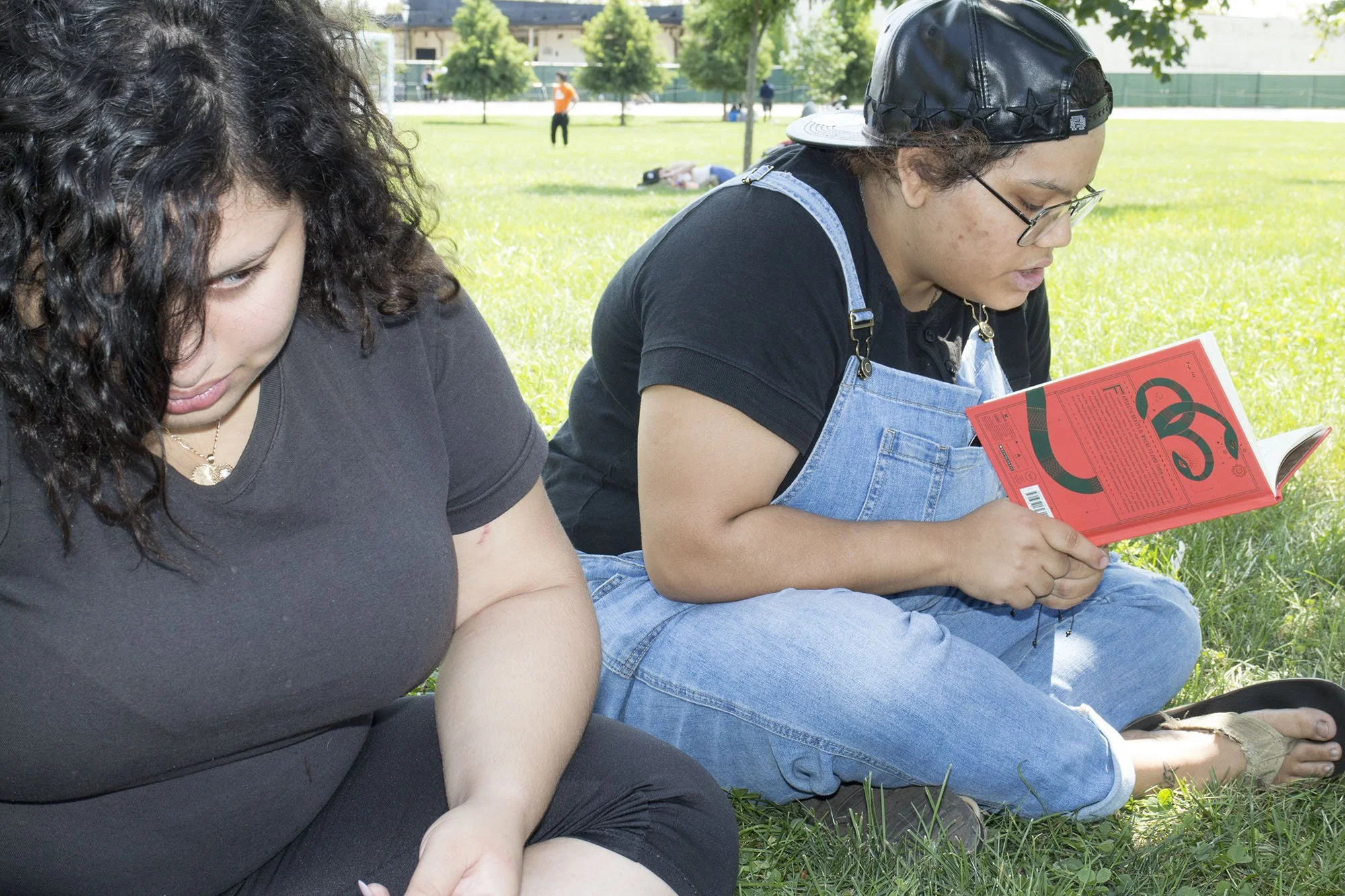 Two women sitting on grass under a tree, one reading a magazine and the other looking down.