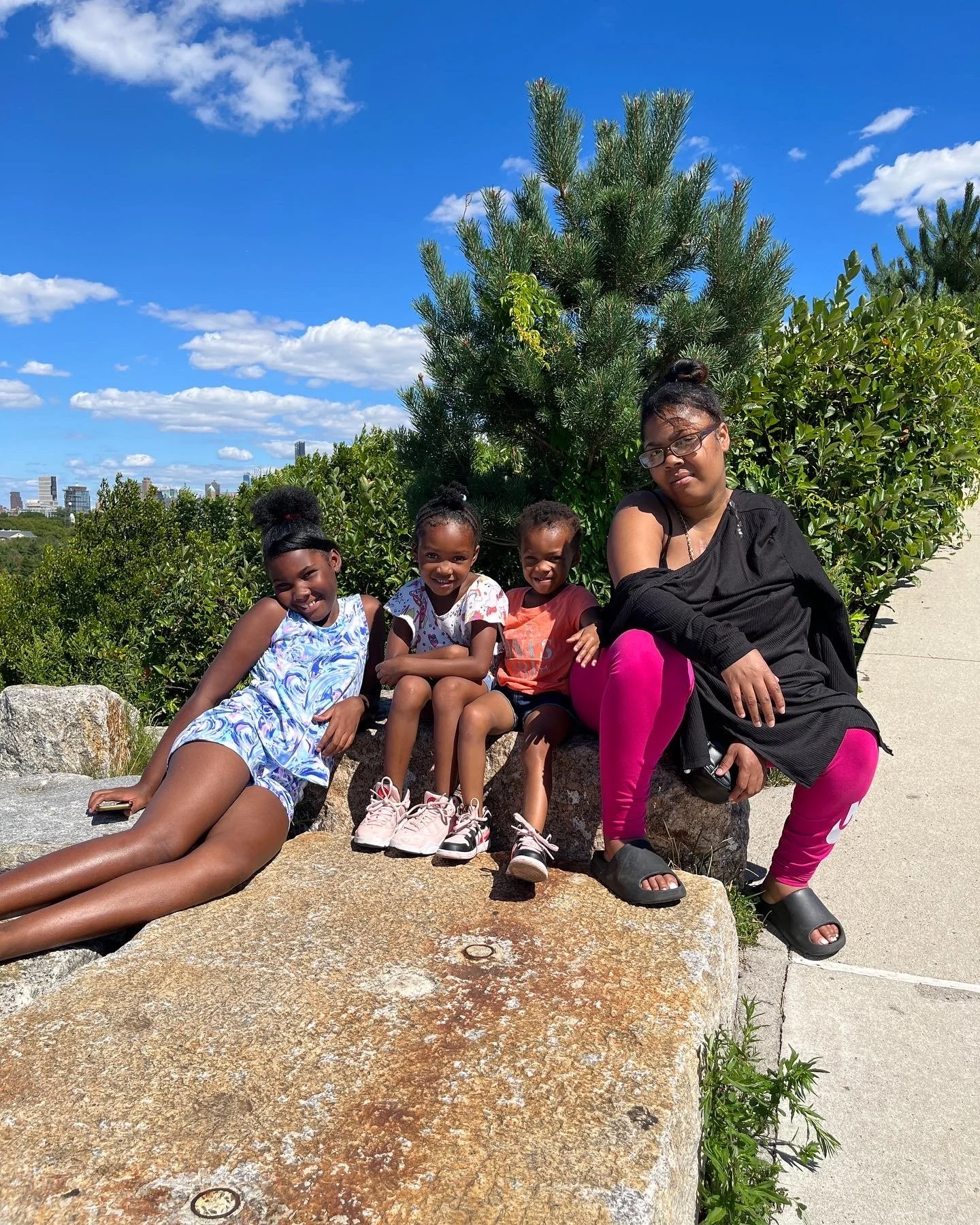 A woman and four young girls sitting on a large rock outdoors on a sunny day with a blue sky and clouds, greenery, and city buildings in the background.
