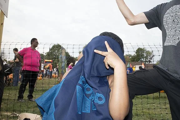Child with a blue hoodie covering their face, sitting outdoors near a wire fence at a park or fair with other people and inflatable structures in the background.
