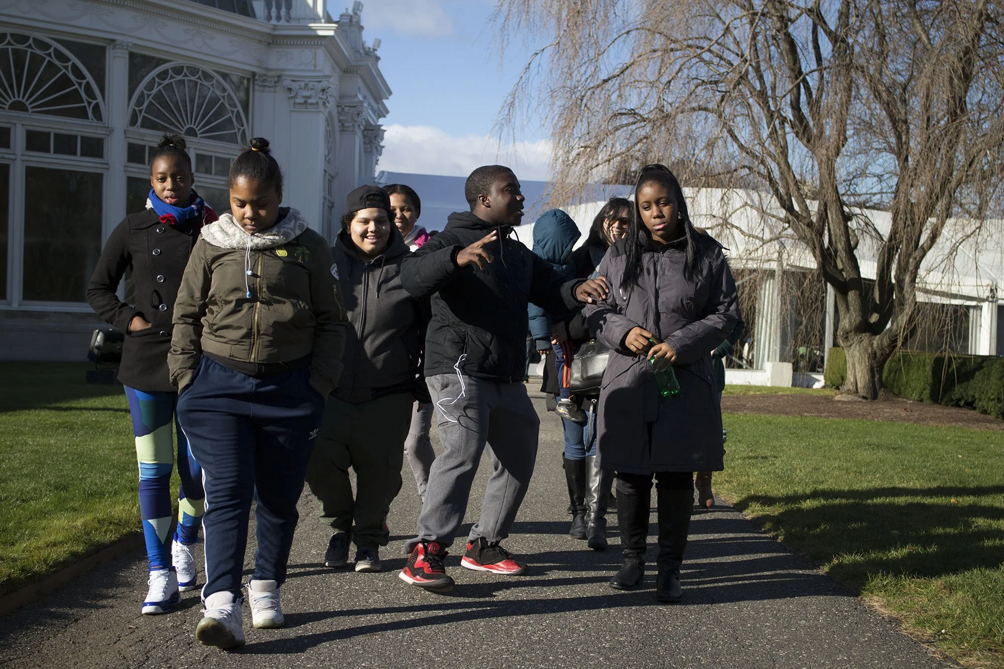 A group of diverse young adults walking outdoors on a sunny day, talking and smiling, with a white building and leafless trees in the background.