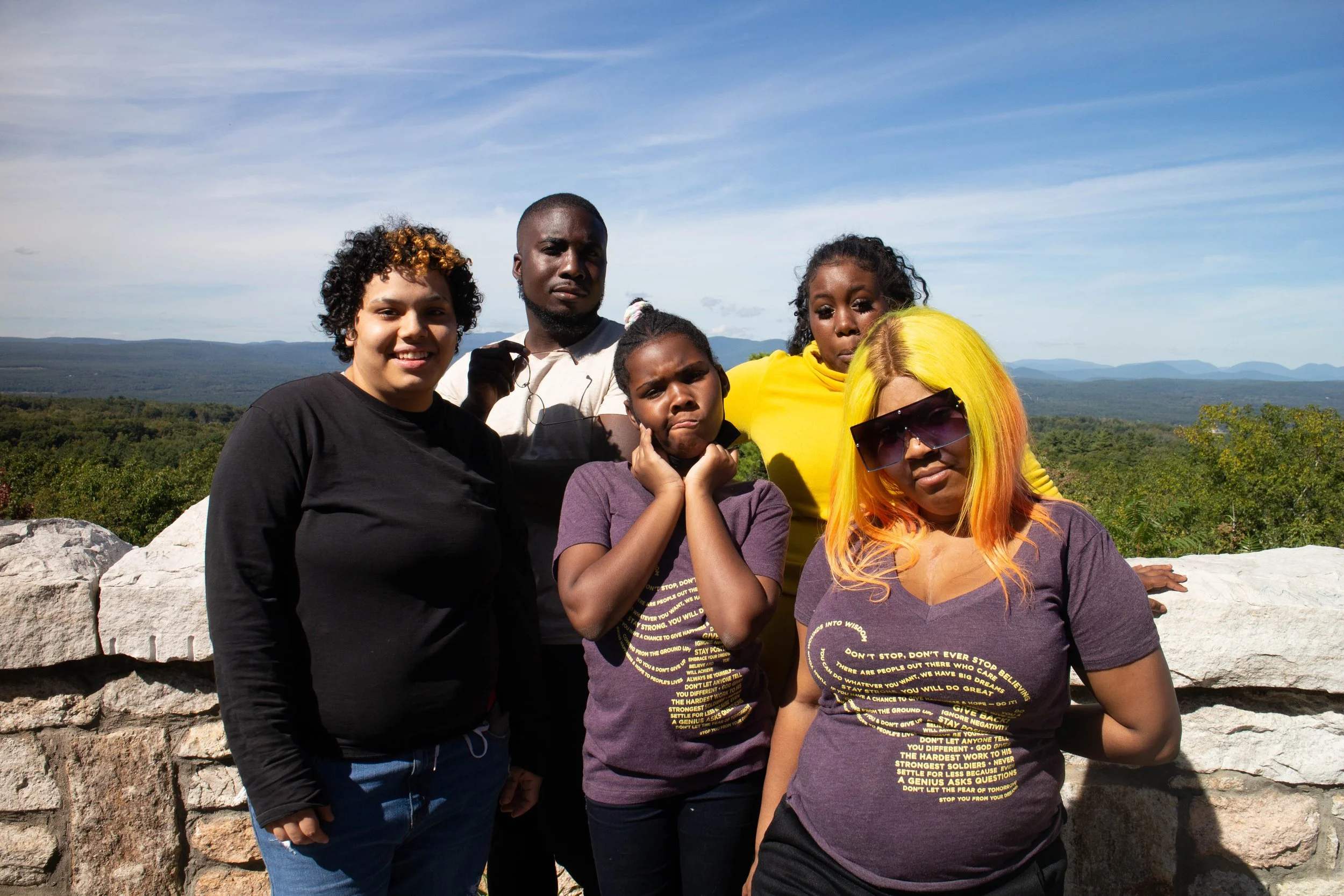 Group of six diverse people outdoors on a stone overlook with mountains and blue sky in the background.