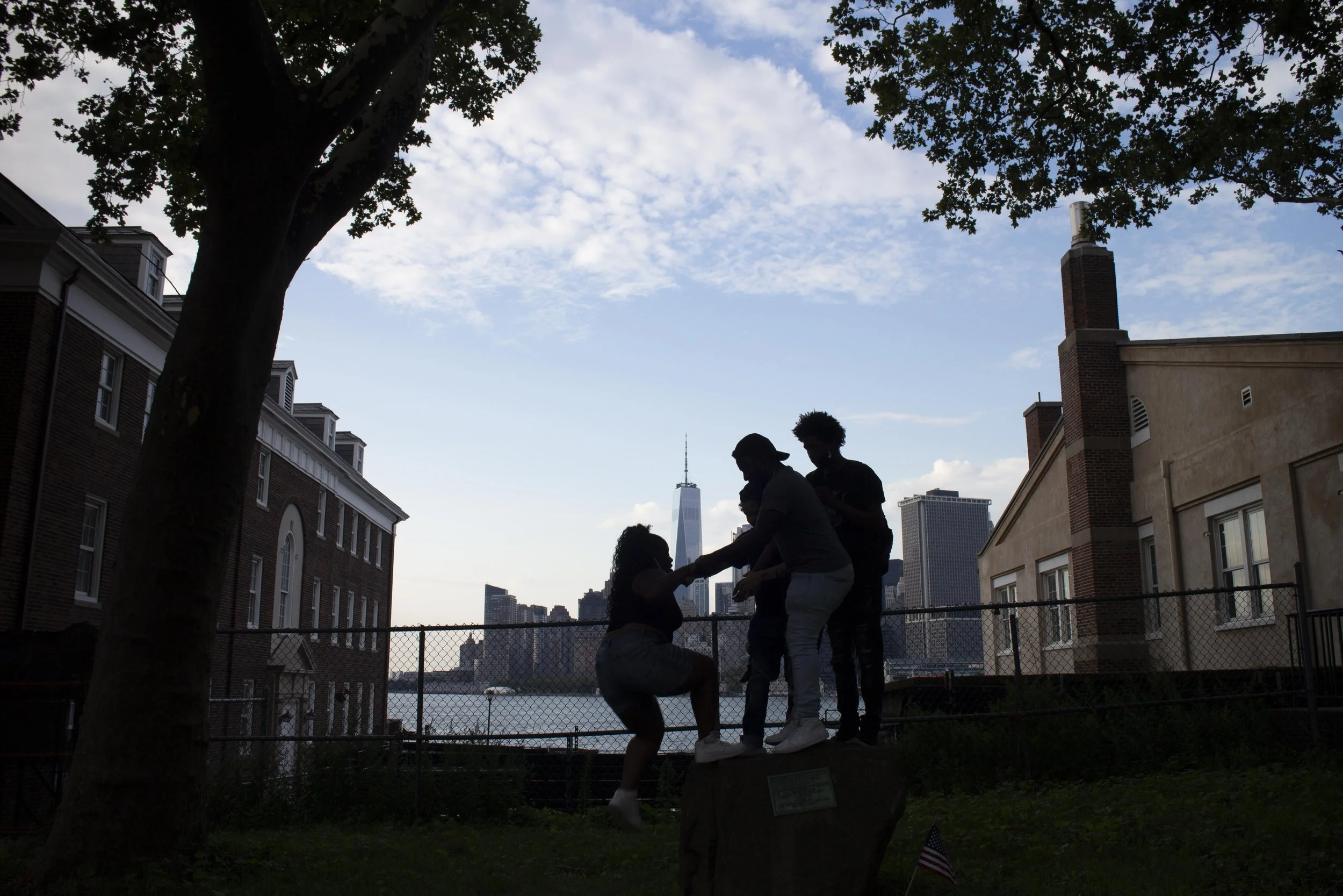 Silhouettes of people standing on a ledge near a chain-link fence, with the New York City skyline and the Statue of Liberty in the background, under a partly cloudy sky, with a large tree on the left side.
