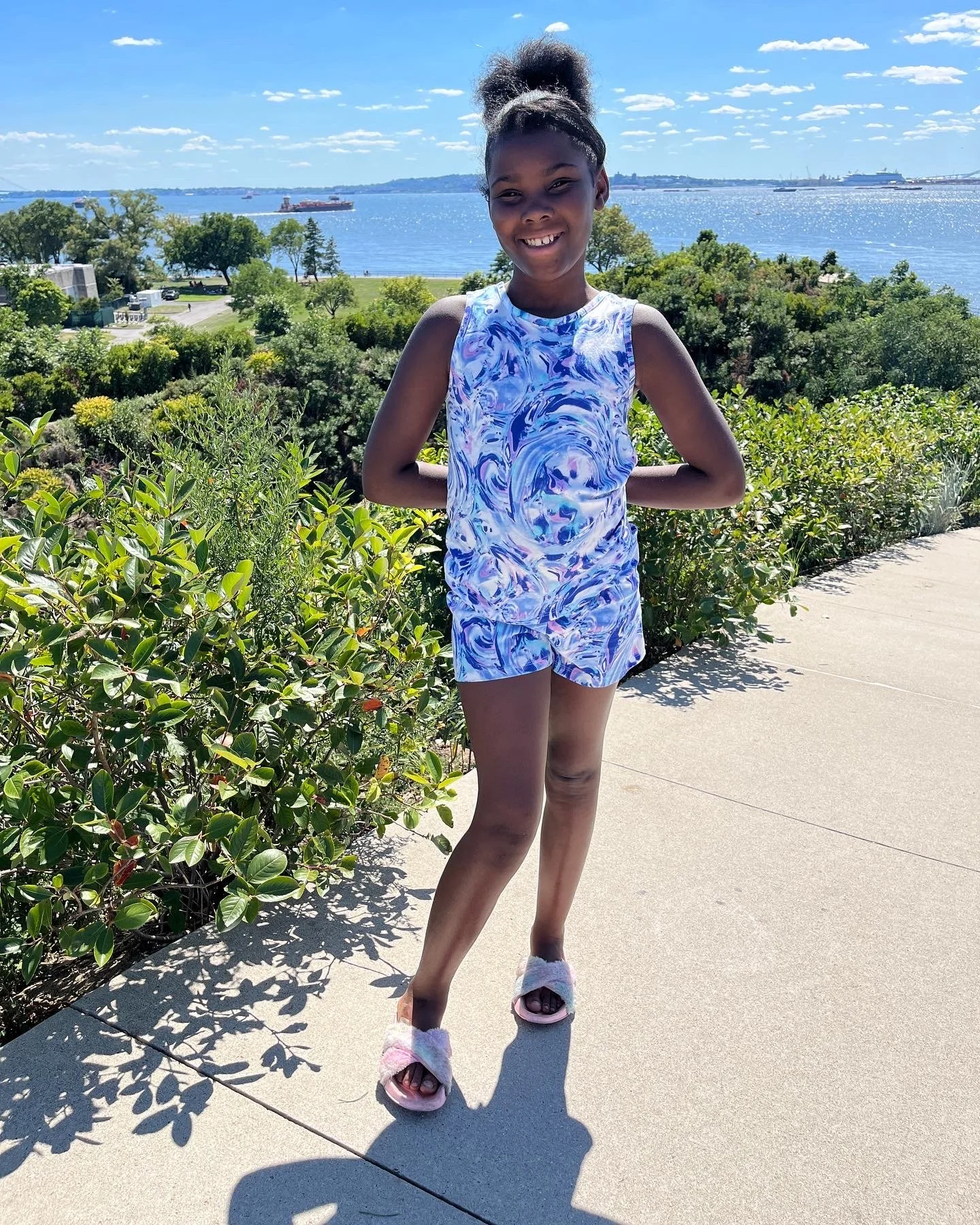 A young girl smiling in a blue and white marble-patterned outfit, standing on a sidewalk with bushes and trees, a body of water, boats, and ships in the background on a sunny day.