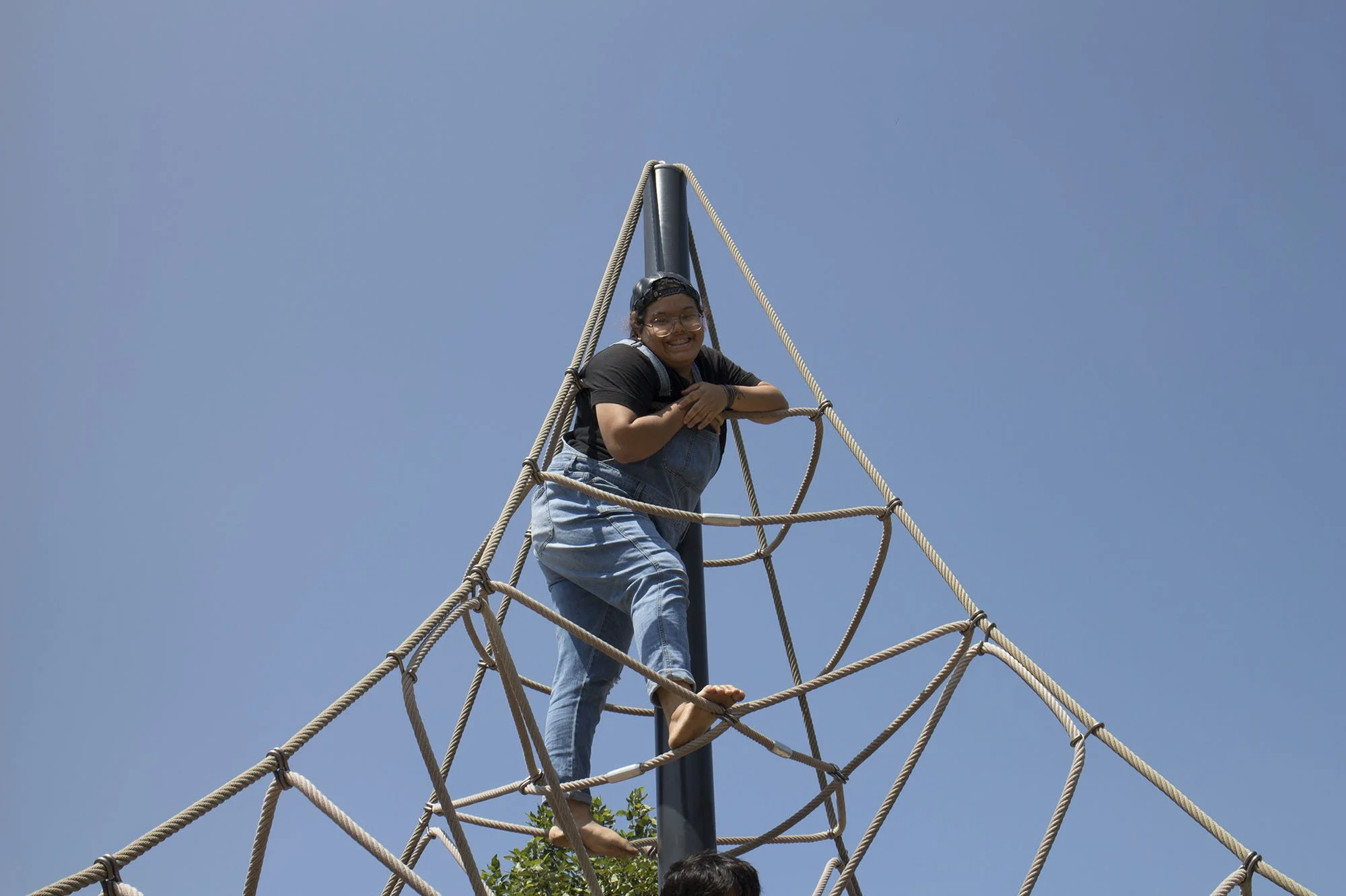 A person smiling and climbing a rope web structure outdoors against a clear blue sky.