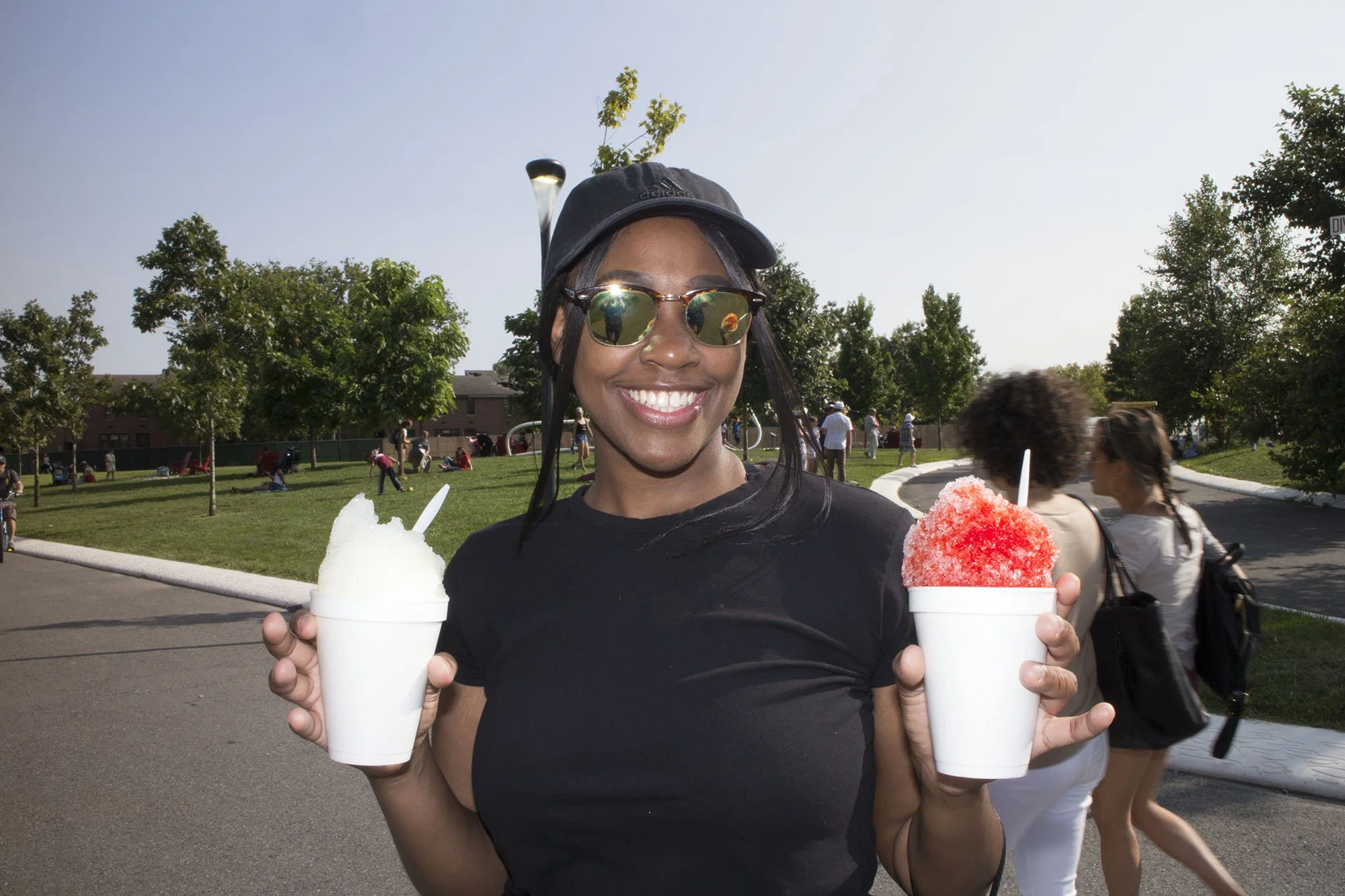Woman wearing sunglasses and a cap smiling and holding two cups of shaved ice, one red and one white, in a park with trees and people in the background.