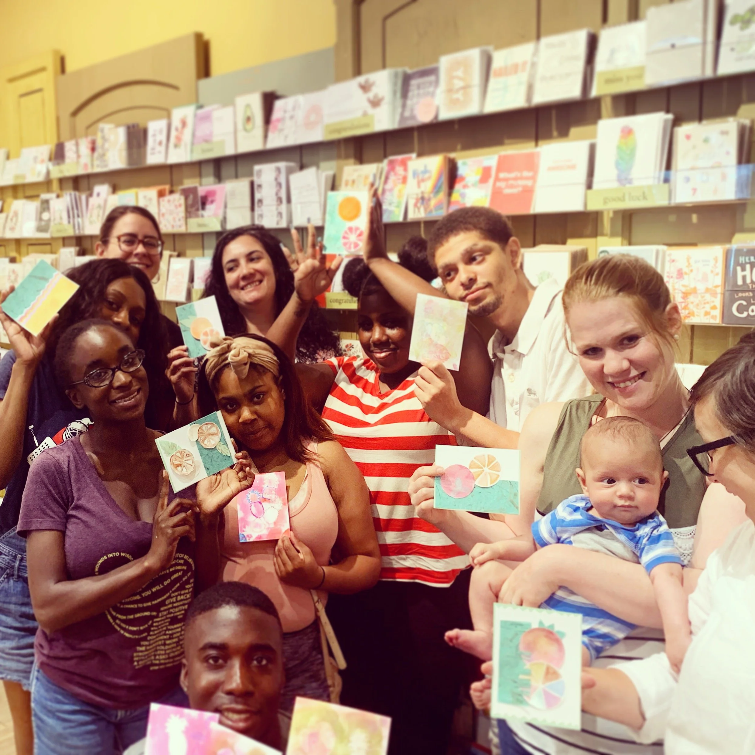 Group of diverse people at a store or event, holding colorful handmade greeting cards, with books displayed on the wall behind them.