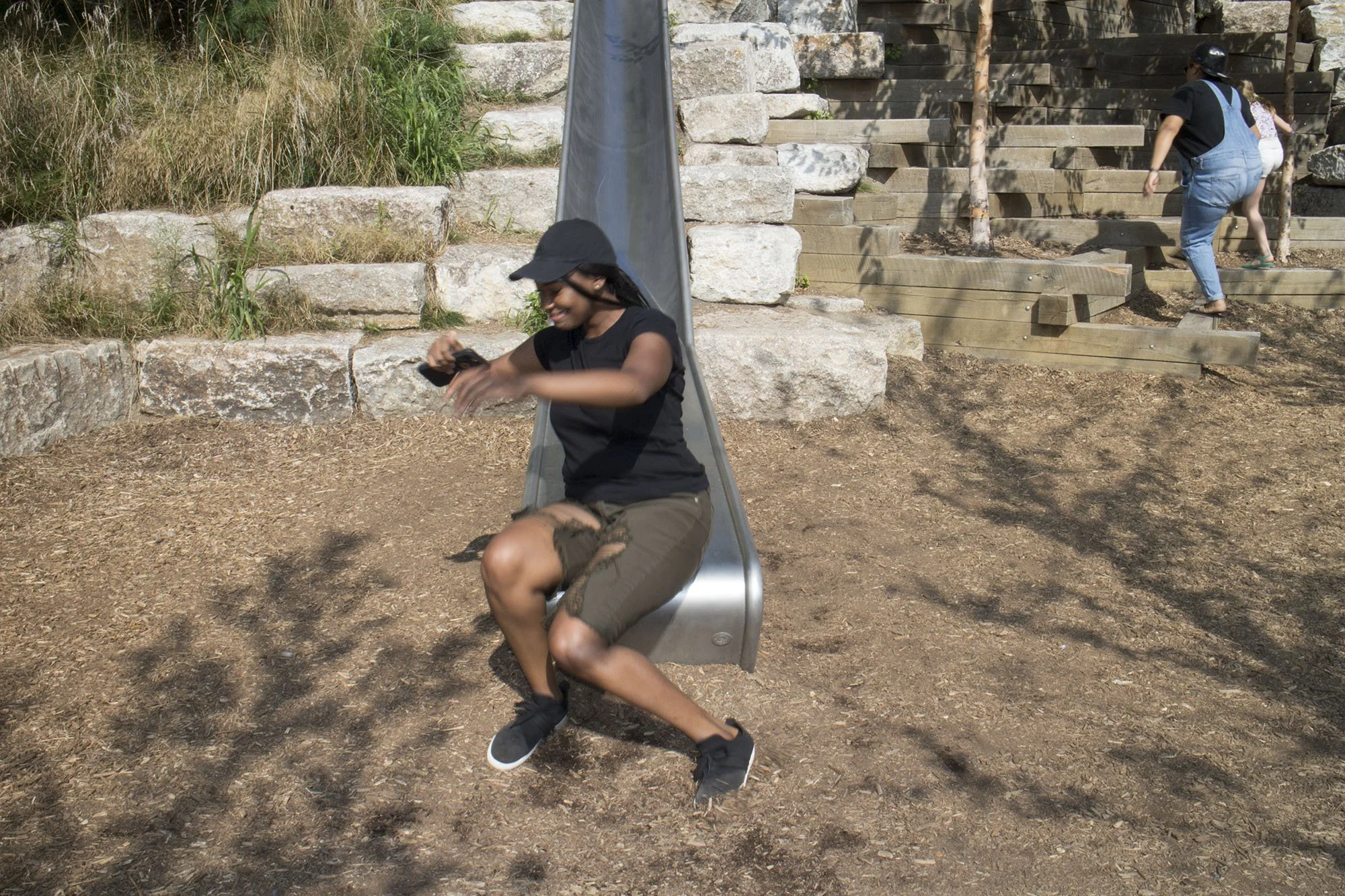 A woman is sitting on a metal slide at a park, holding a phone and smiling. In the background, two children are climbing stone and wooden steps on a hillside.