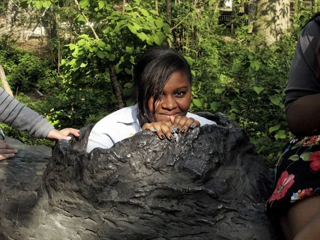 Girl with medium skin and dark hair leaning over a large black rock, outdoors with green foliage in the background.