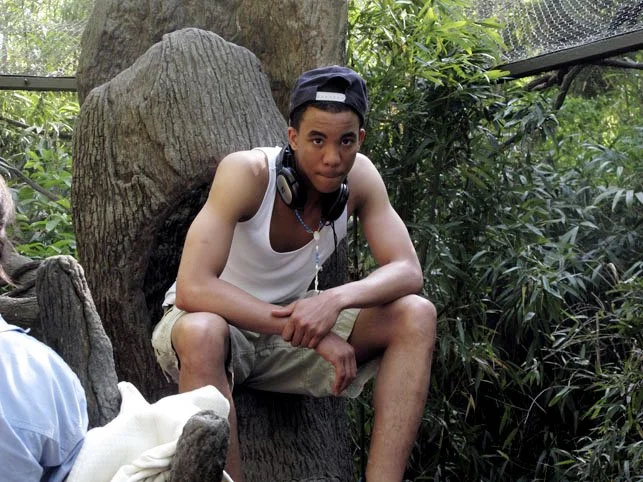 Young man sitting on a large tree trunk in a lush, green outdoor setting with headphones around his neck.