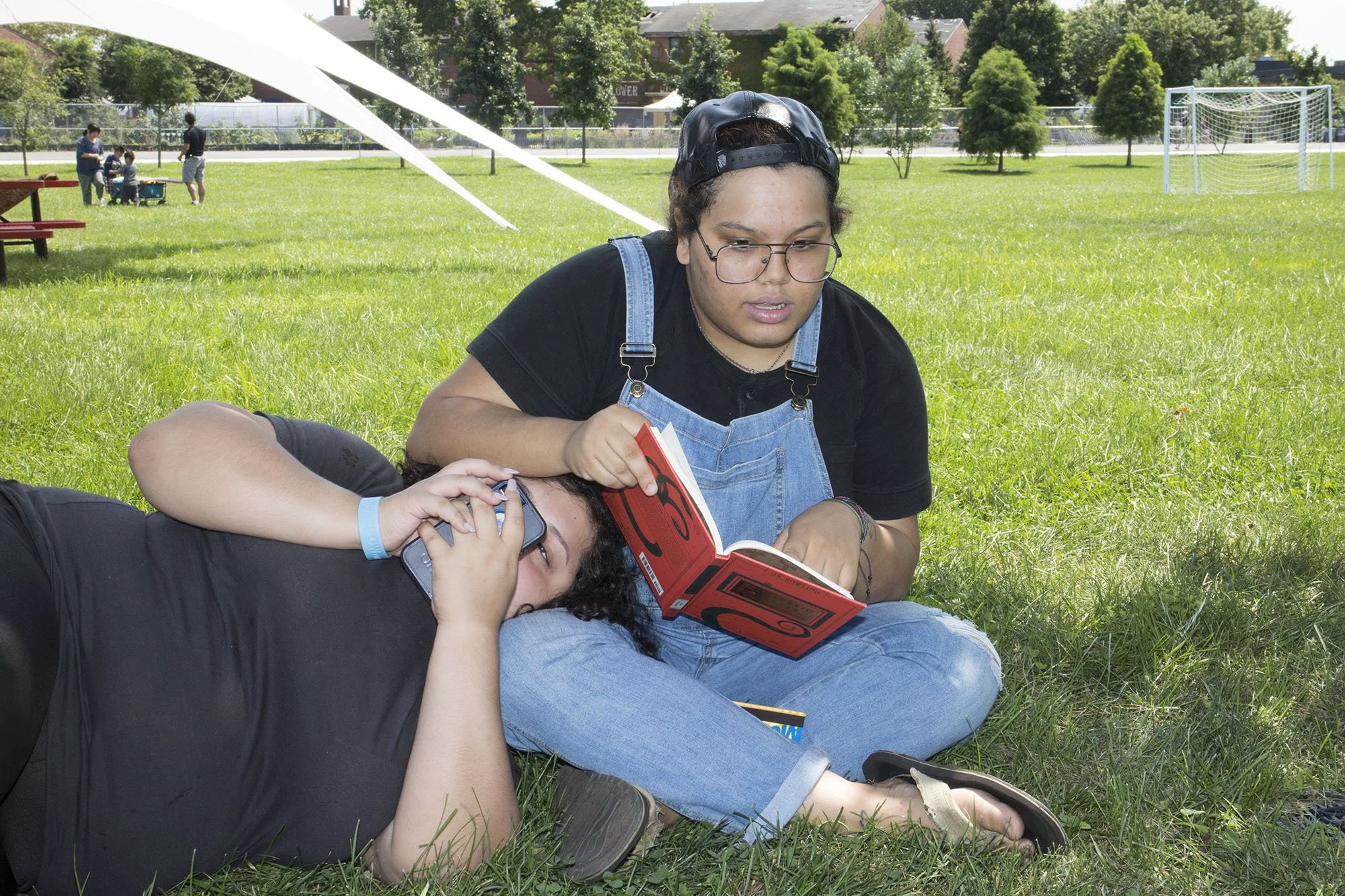Two teenage girls are lying on the grass in a park, one talking on a phone and the other reading a book. In the background, there are children playing near a group of picnic tables and a soccer goal.