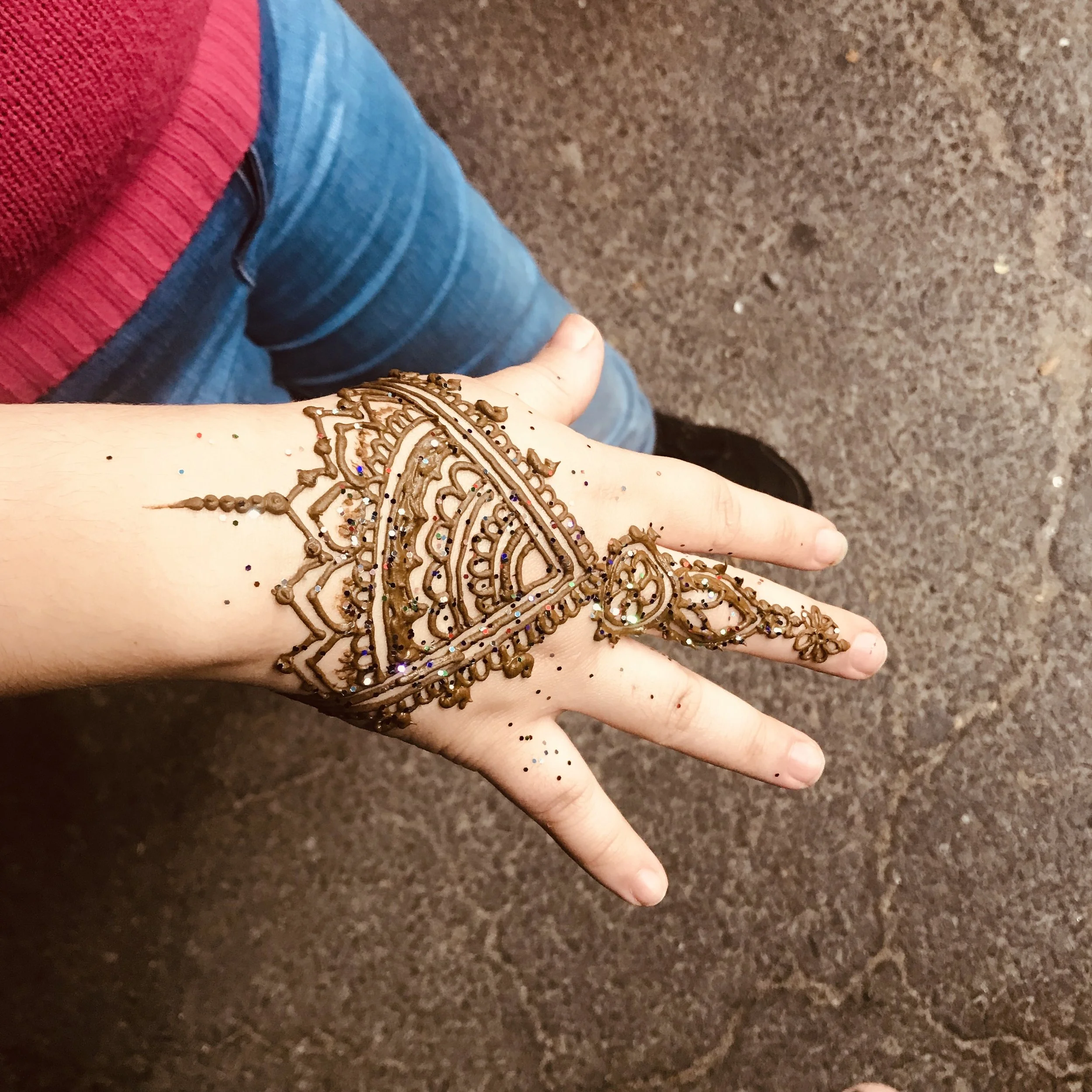 Hand decorated with intricate henna design featuring a large semi-circle pattern and small embellishments, resting on a surface with part of a person's arm and clothing visible.