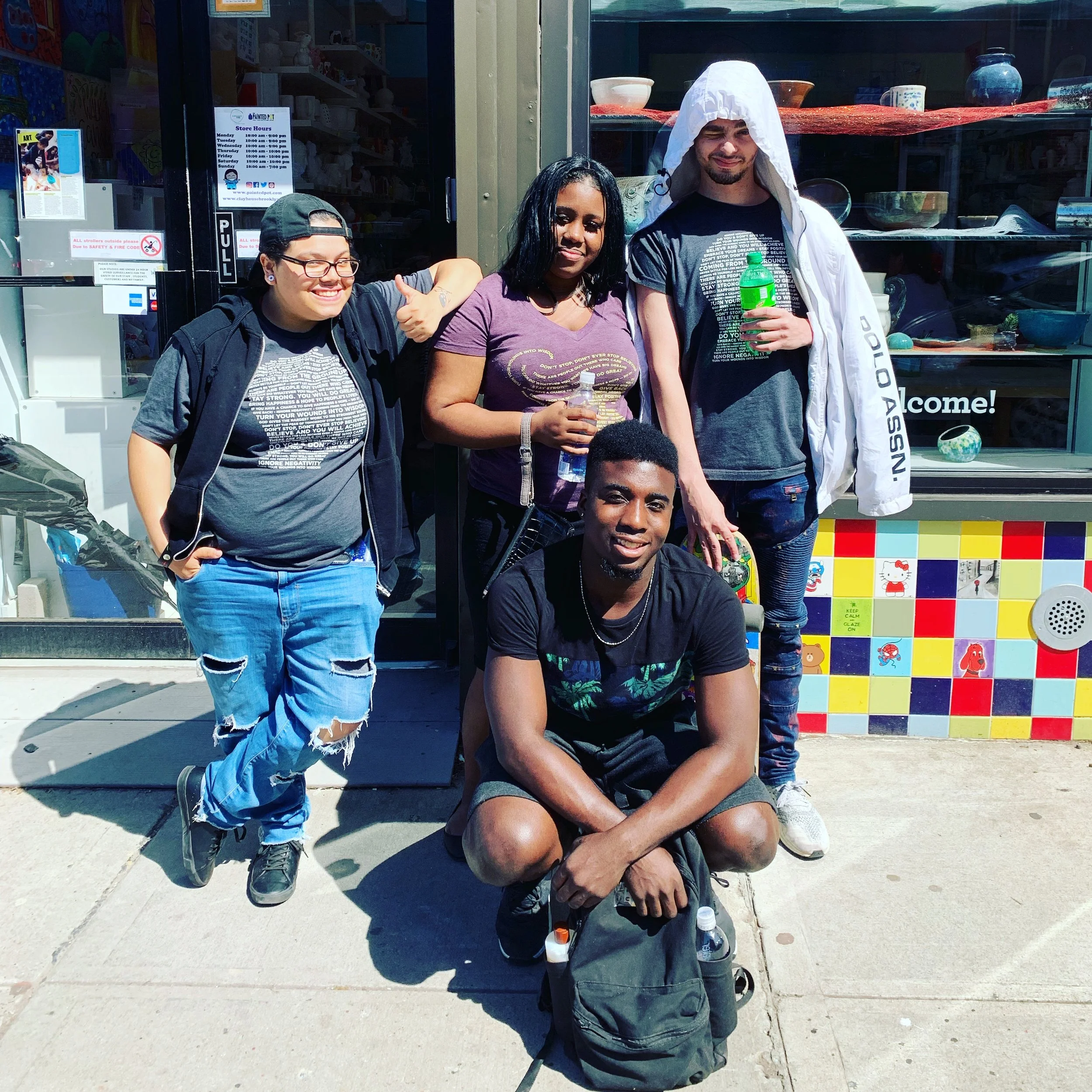 Group of five young adults posing outside a storefront with colorful tiles and various displayed items. They are smiling and casually dressed, with some holding bottled drinks.