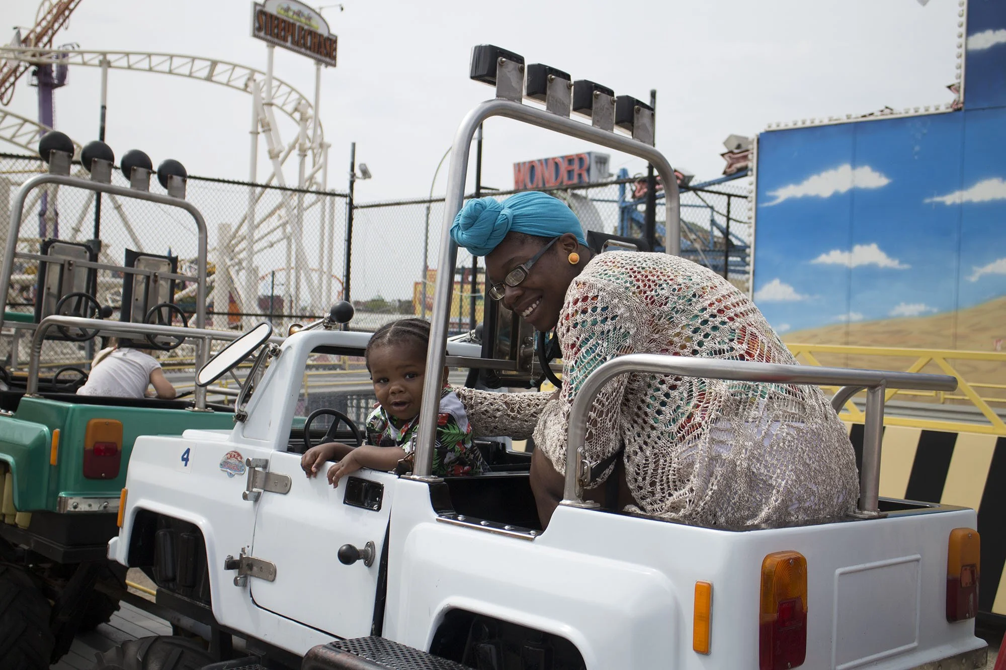 A woman with glasses, earrings, and a blue headscarf smiling in a white toy car ride at an amusement park with a child inside, with a backdrop of amusement park rides and a painted sky mural.
