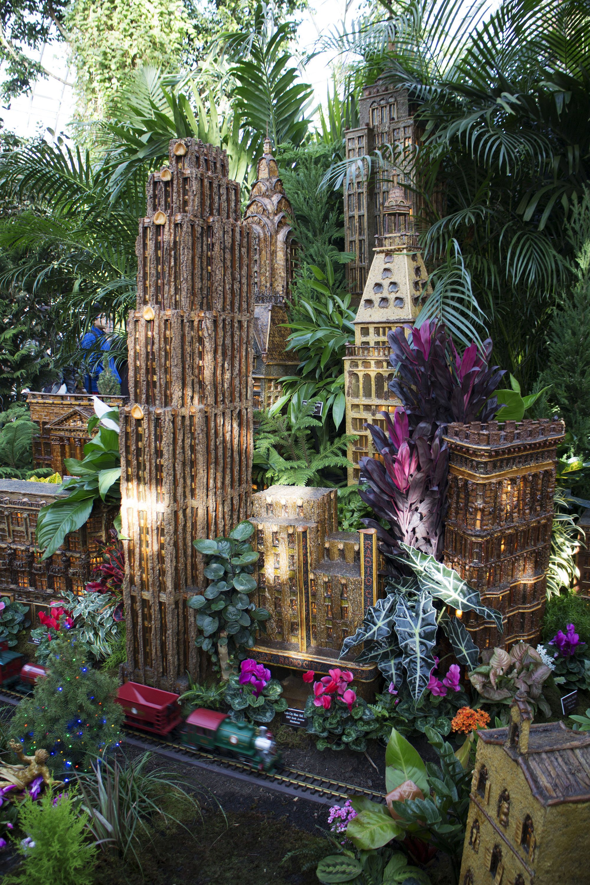 Miniature model of a cityscape with tall buildings, surrounded by lush green plants, flowers, and a small toy train on tracks.