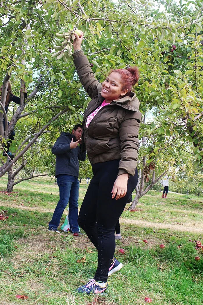 A woman smiling and reaching for an apple in an orchard, with a man behind her giving a thumbs-up."}