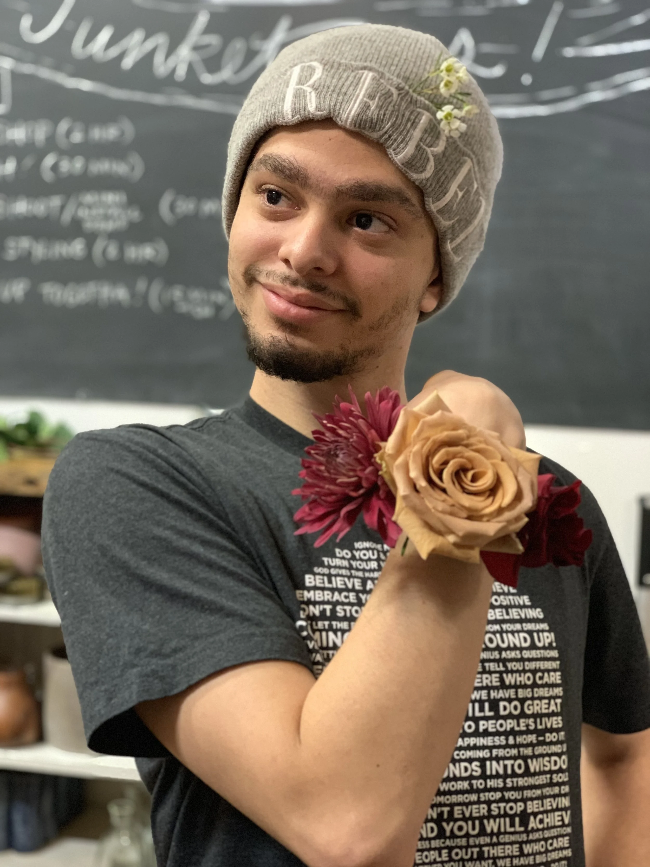 A young man with a goatee wearing a knit gray beanie with flowers and a black T-shirt, holding a bouquet of flowers close to his chest in an indoor setting with a chalkboard in the background.