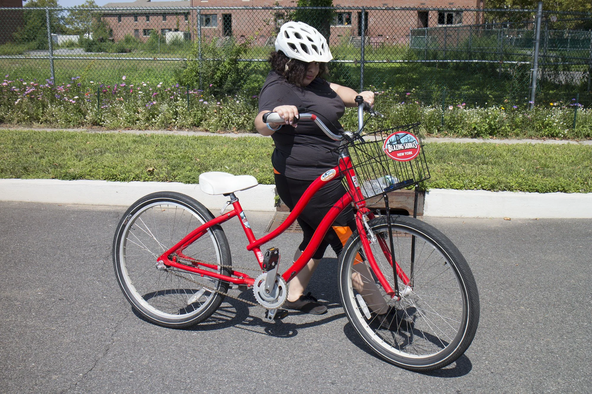 Person with curly hair wearing a white helmet walking with a red bicycle on a paved road, with a flower bed and a chain-link fence in the background.