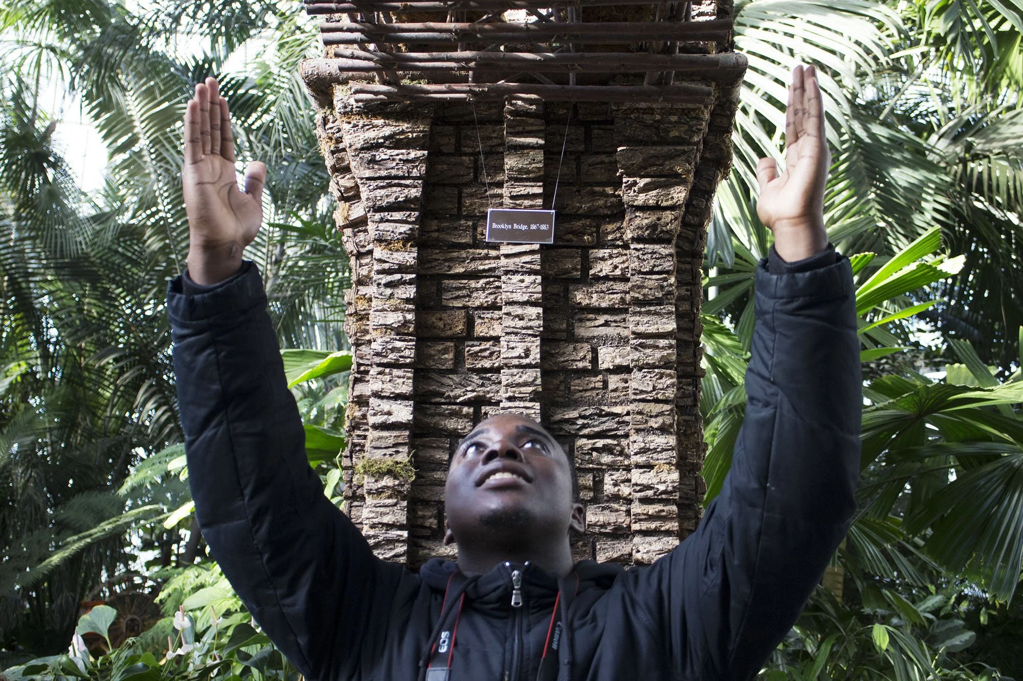 A person with a black jacket looking up and raising both hands towards a miniature model of Brooklyn Bridge, surrounded by lush green foliage.