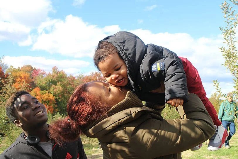 A woman holding a young boy in a park with autumn trees in the background, while a man looks on and smiles nearby.