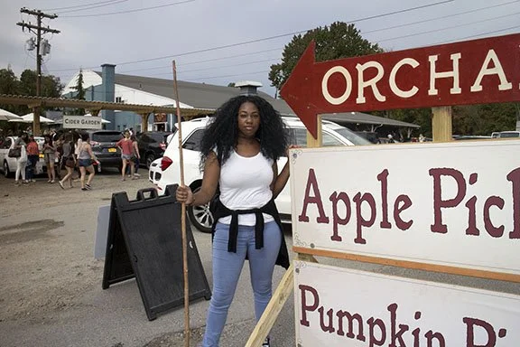 Young woman standing next to signs for apple picking and pumpkin picking at an orchard during daytime.