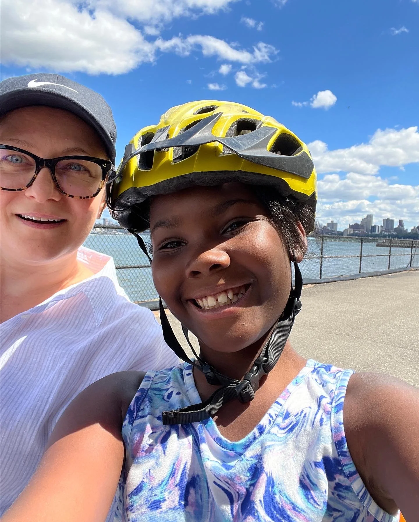 A smiling woman and girl wearing bike helmets taking a selfie outdoors near a body of water with a city skyline in the background on a sunny day with some clouds.