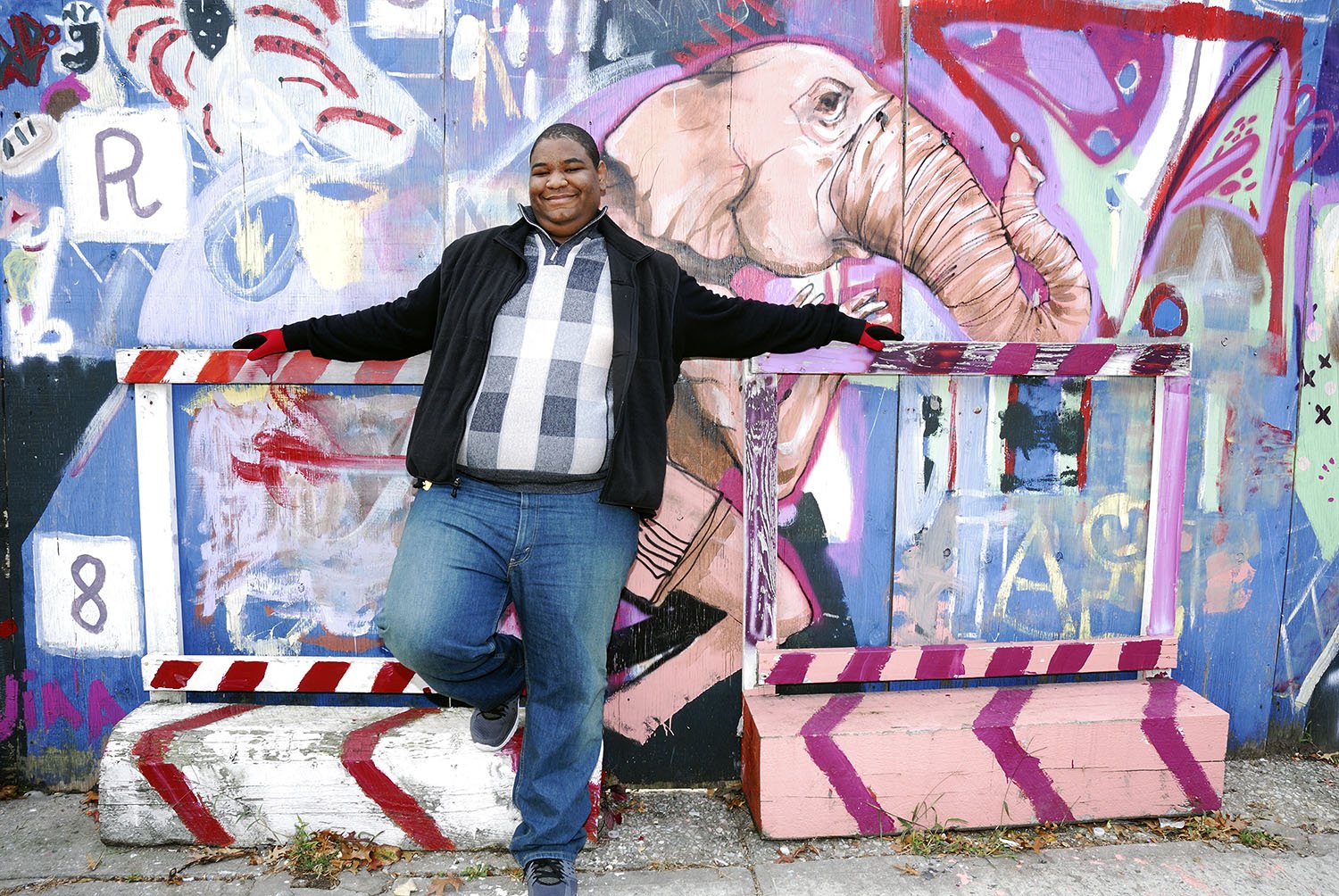 A man smiling and posing with arms outstretched in front of colorful street art featuring an elephant and abstract designs.
