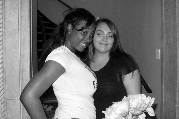 Two women standing close together, smiling, with one holding a bouquet of flowers, in a black and white photo indoors.