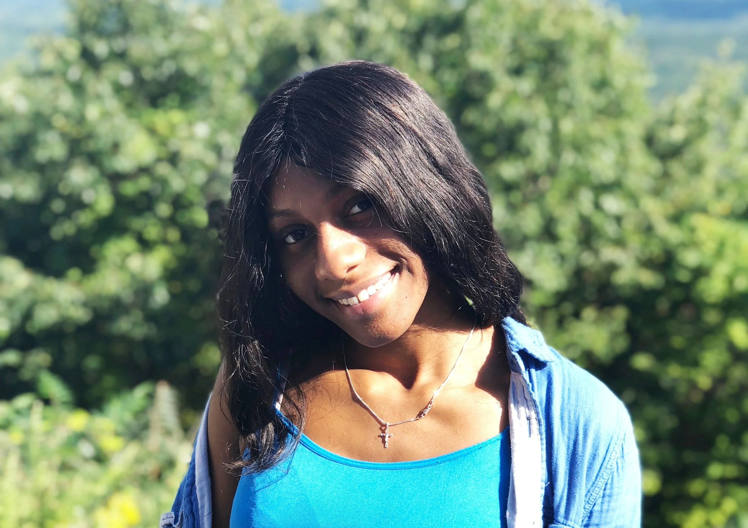 A woman with shoulder-length black hair wearing a blue top and necklace outdoors with green trees in the background.