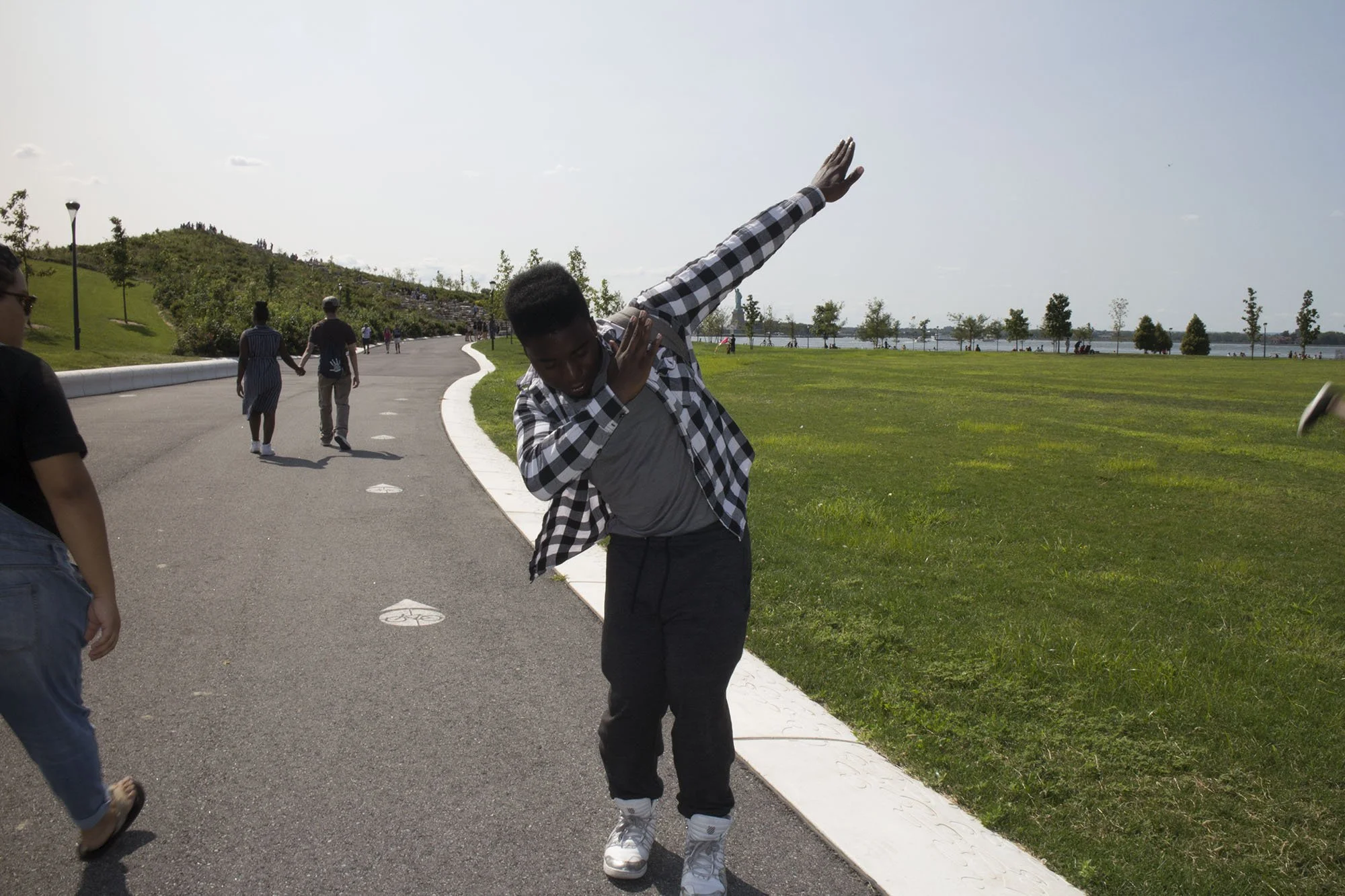 A person performing a dance move outdoors on a paved path near a grassy area and a body of water, with other people walking in the background.