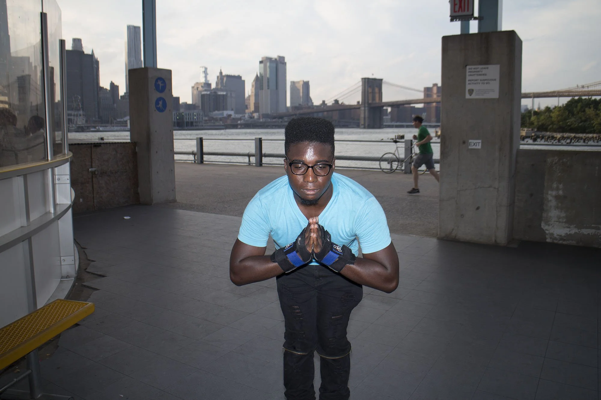 A young man wearing glasses and a light blue t-shirt stands with his hands pressed together in a prayer or meditation pose, outdoors near a waterfront with a city skyline and Brooklyn Bridge in the background.