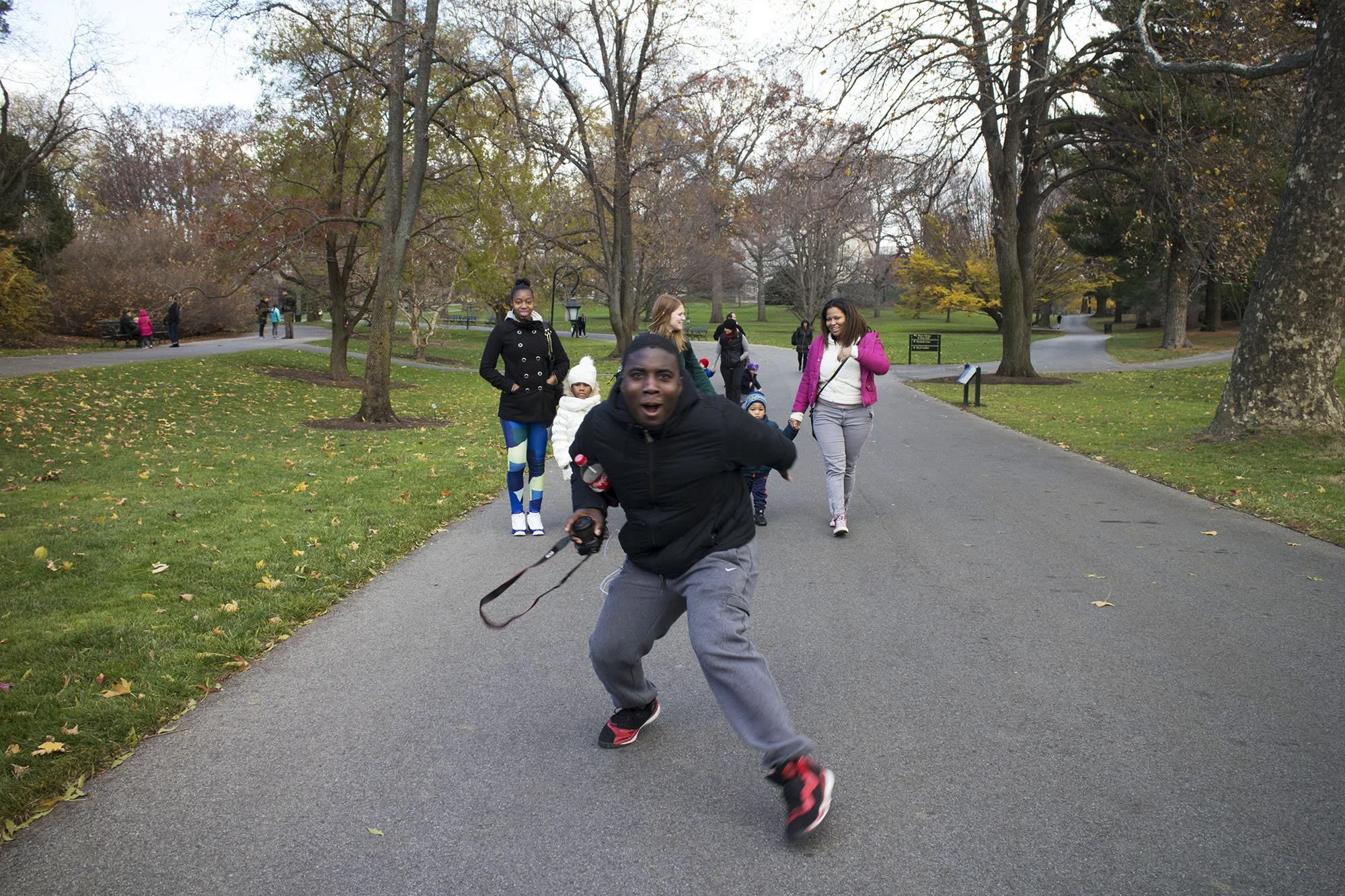Children and adults enjoying a walk in a park during autumn, with trees shedding leaves and a paved path.